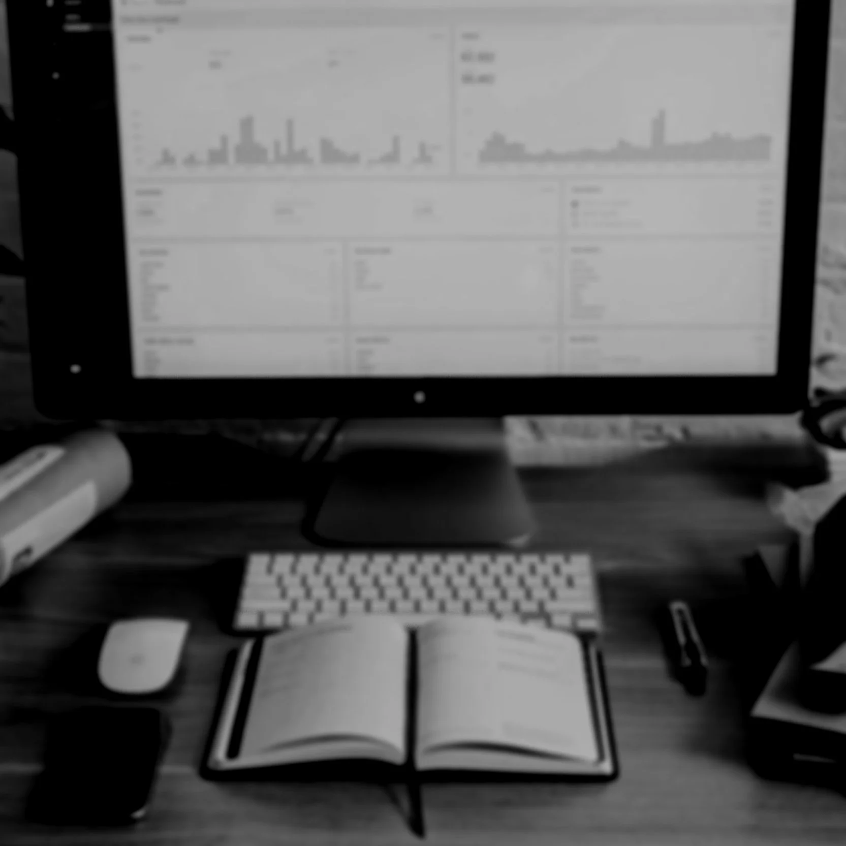 Black and white photo of a computer monitor displaying charts and graphs, a keyboard, mouse, open notebook, pen, and various office supplies on a wooden desk.