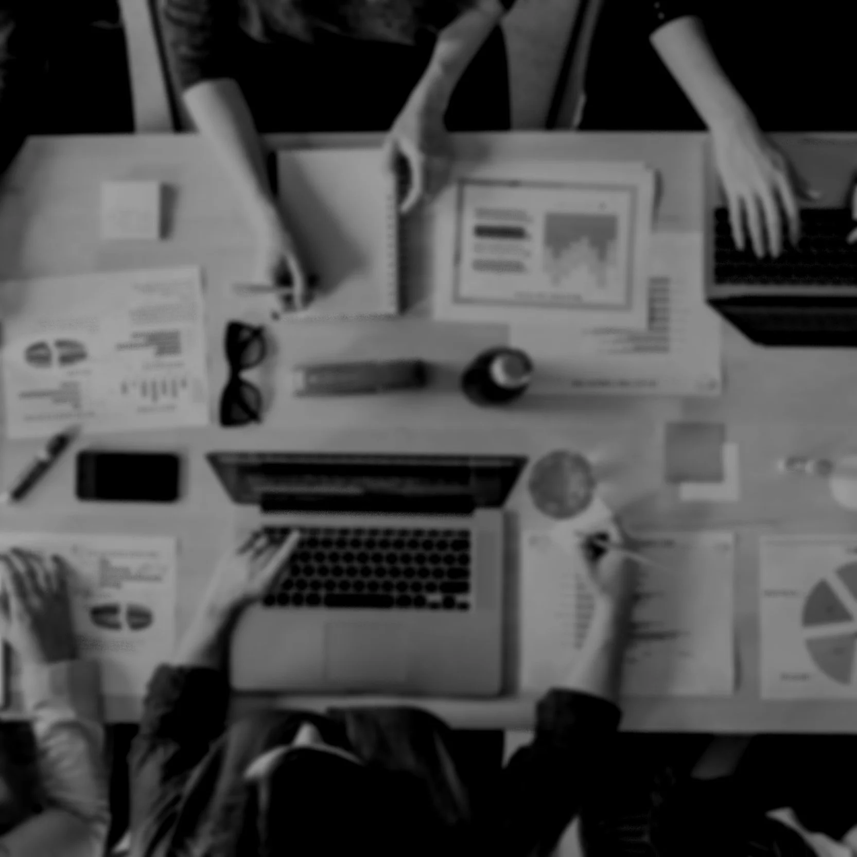 Overhead view of a cluttered desk with papers, a laptop, glasses, a pen, a cylindrical container, and people working or collaborating.