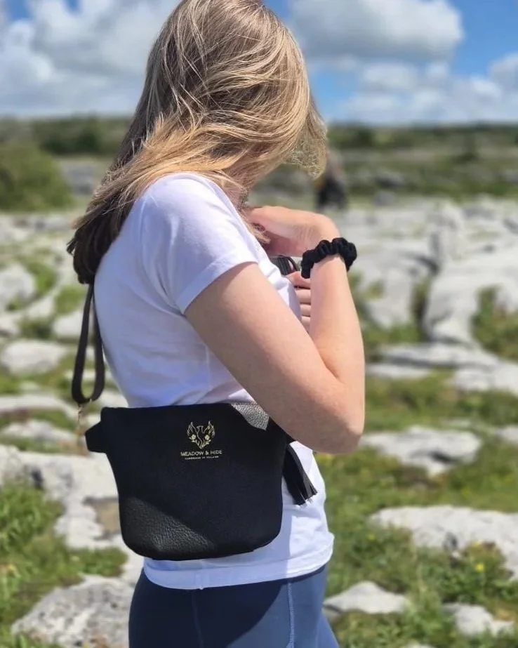 A woman with blonde hair in a white T-shirt standing outdoors in a rocky, grassy landscape in the Burren in County Clare. She has a dark olive crossbody bag with a logo that reads 'Meadow & Hide' on it.