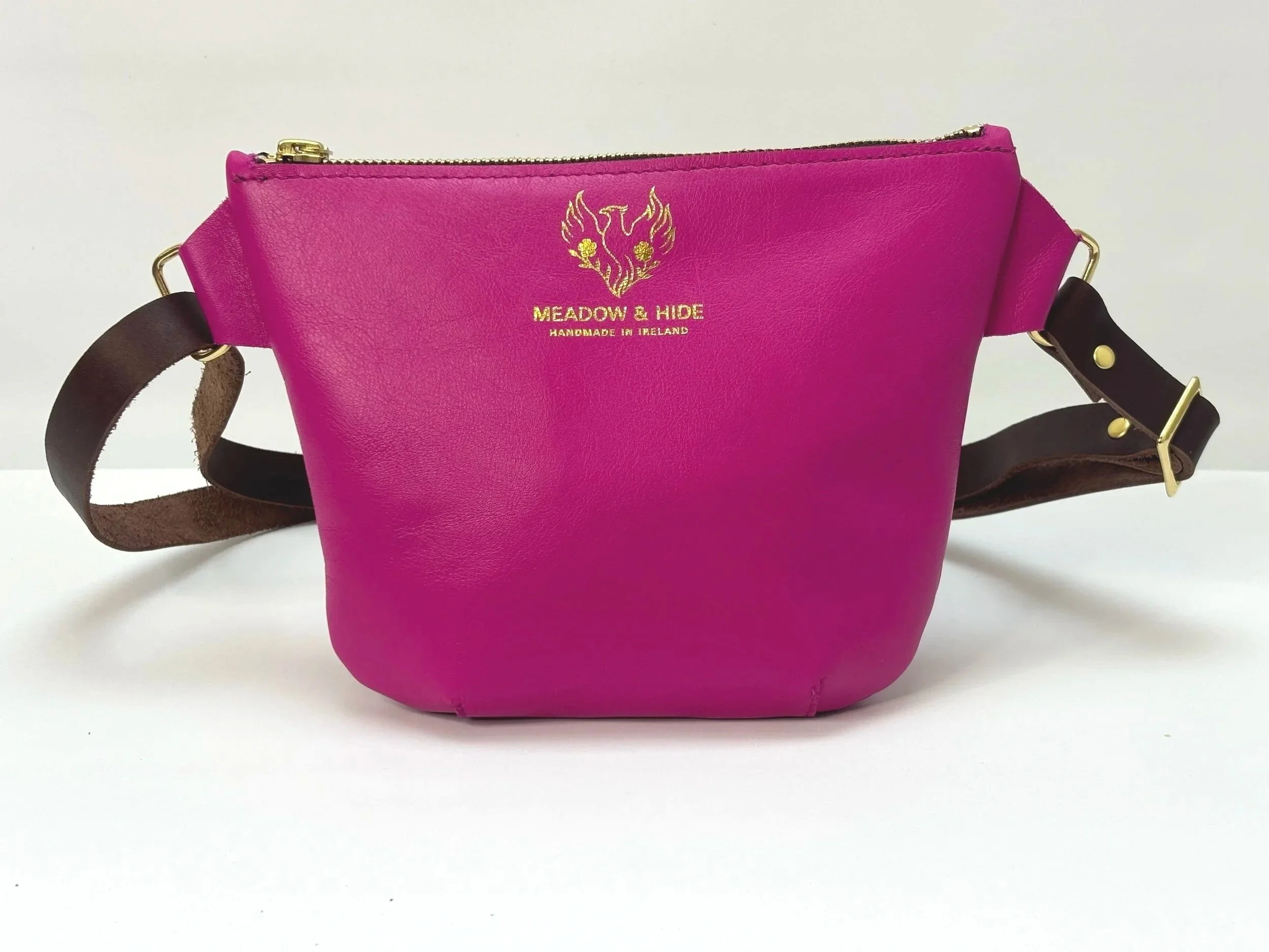 Pink leather handbag with a gold zipper and a black adjustable strap, featuring gold printed text that reads 'Meadow & Hide Handmade in Ireland' and a gold line drawing of a phoenix.