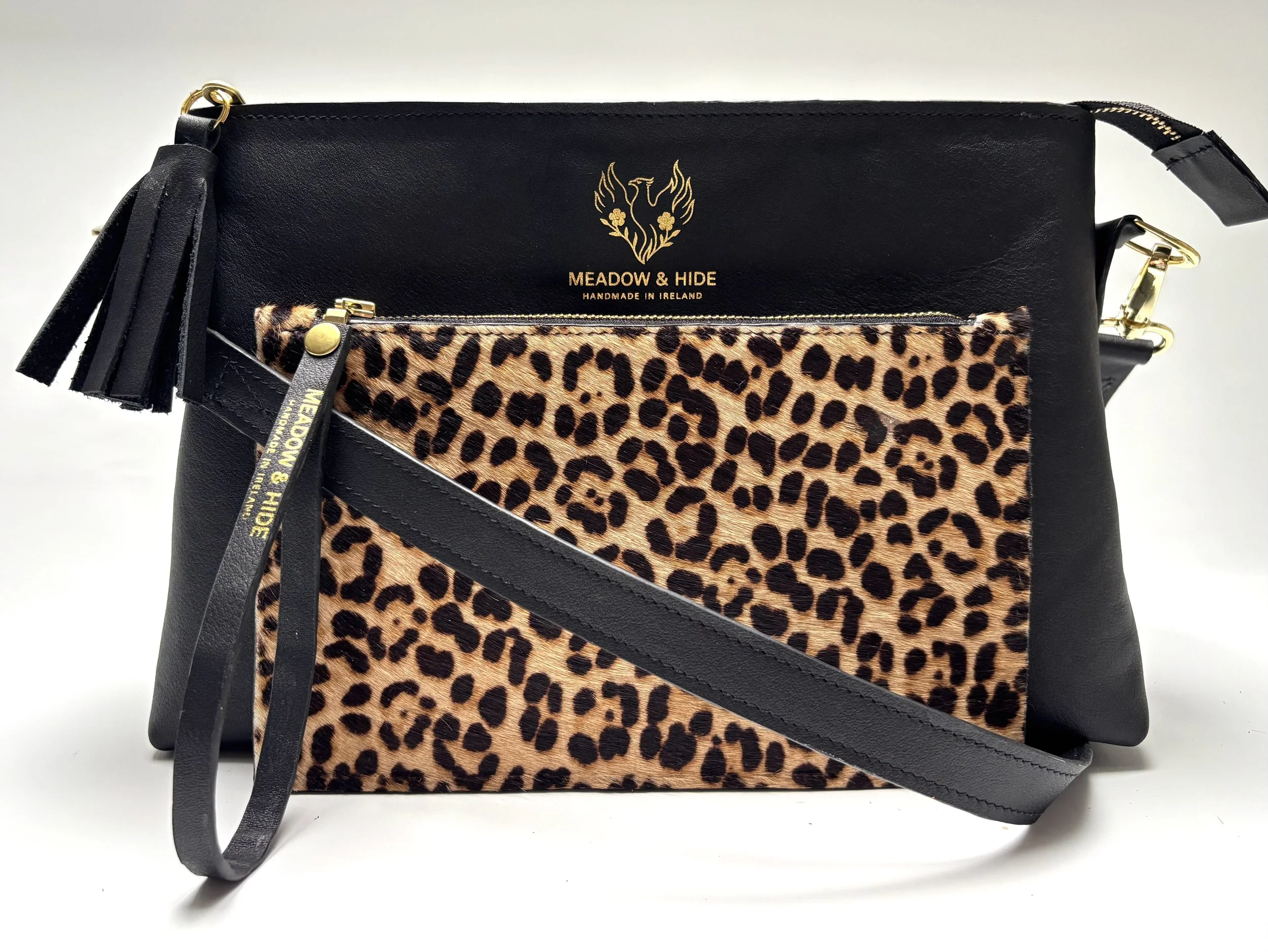 A black handbag with gold hardware, featuring a leopard print fabric pocket, a tassel keychain, and a gold  Meadow and Hide logo with a bird design and the text "Meadow & Hide, Handmade in Ireland".