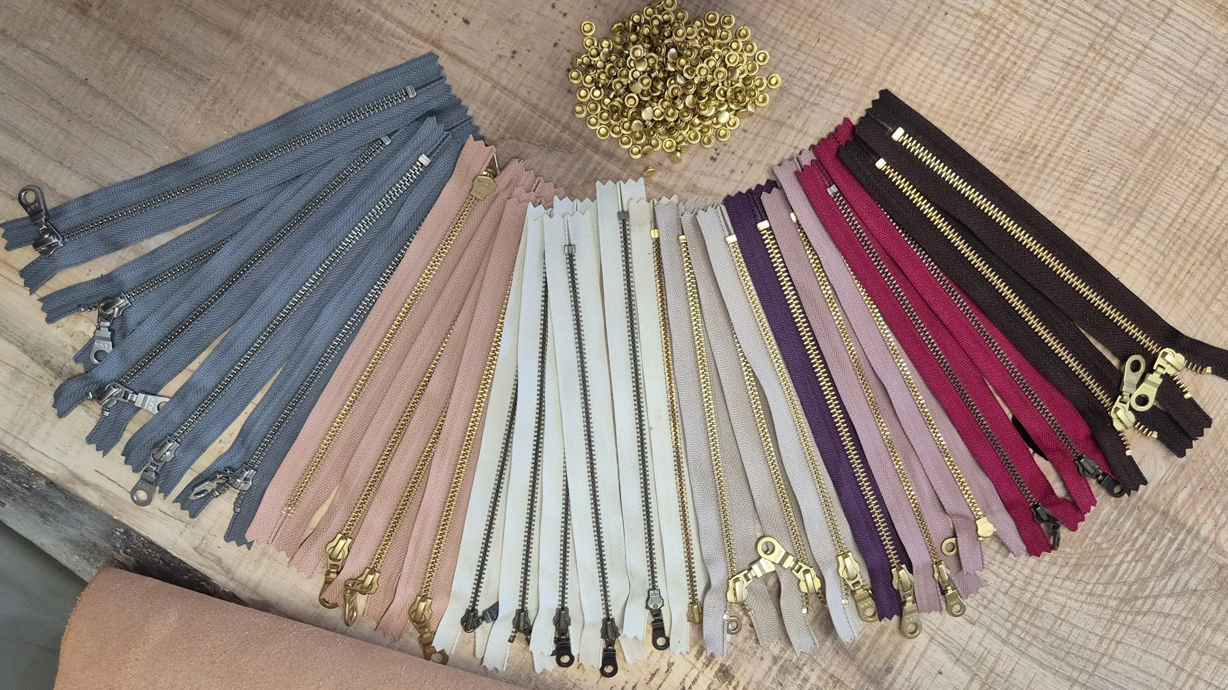 A variety of colorful YKK zippers with gold and silver teeth and pulls spread out on a wooden surface in the Meadow & Hide workshop, along with a small pile of gold-colored rivets