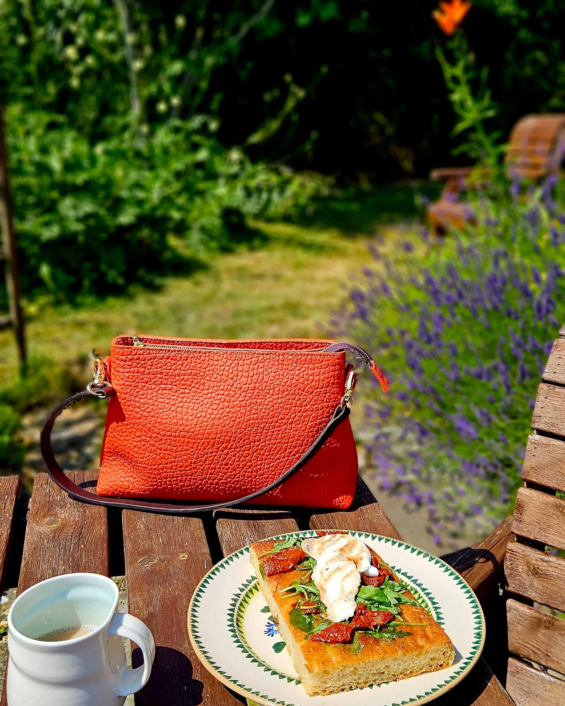 A table set outdoors with focaccia. Next to the plate, there is a Meadow & Hide orange leather handbag on a wooden bench, and two cups . The background features lush greenery and purple lavender flowers.