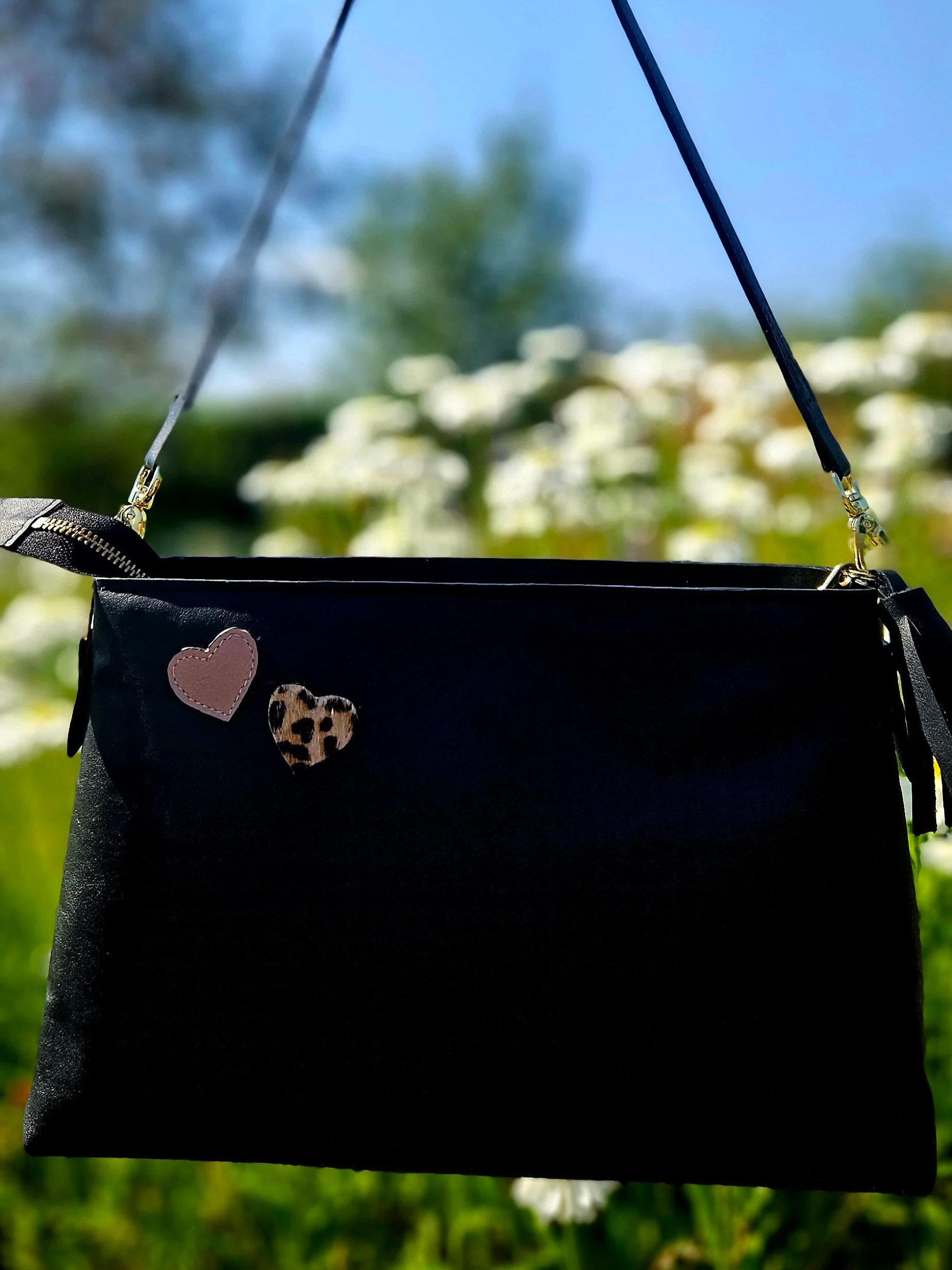 Black bag with heart and leopard print appliques designs, hanging outdoors against blurred greenery and blue sky background.