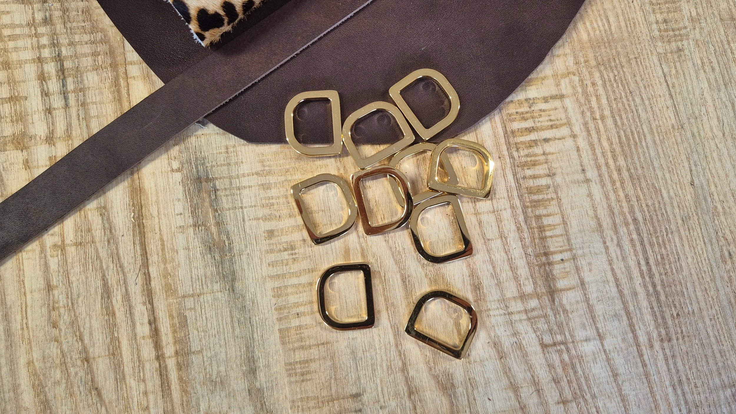 Various D rings scattered on a wooden surface next to a brown leather and leopard print hair on leather in the Meadow & Hide workshop.
