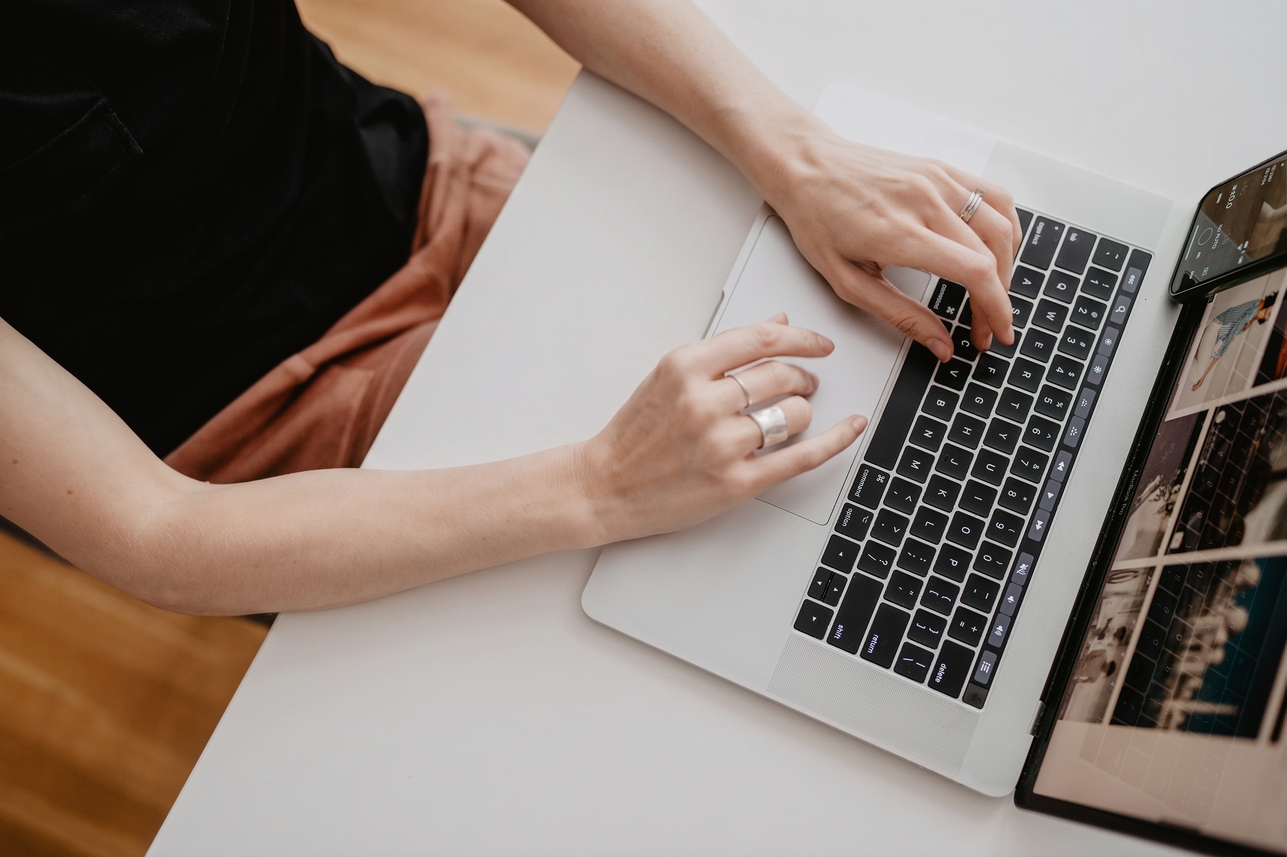 A person using a laptop and smartphone, typing on the keyboard with one hand and holding the phone with the other, on a white desk.