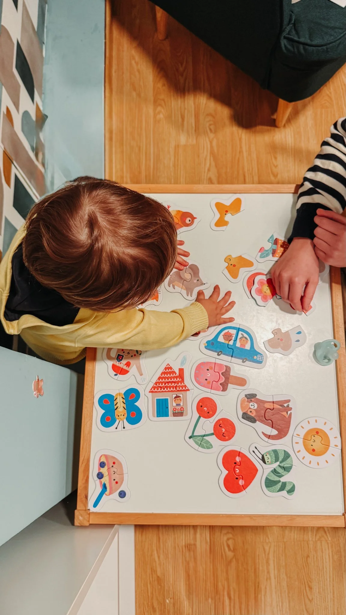 Criança explorando um quebra-cabeça com figuras de animais, casa, carro, e natureza, ao lado de um adulto em um ambiente interno.