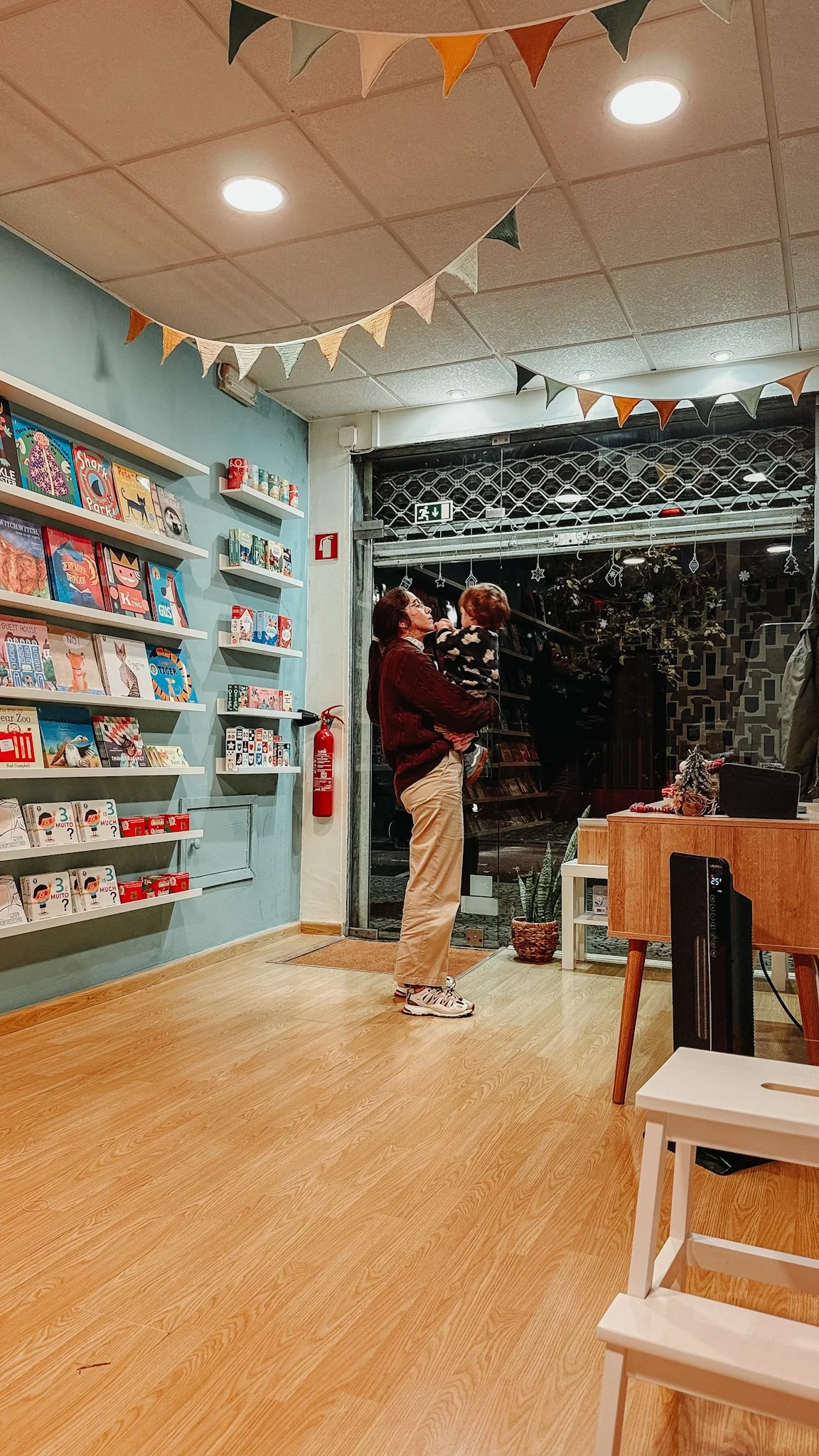 Uma mulher segurando uma criança de cabelo ruivo dentro de uma loja, perto da entrada, com prateleiras de livros na parede ao lado.