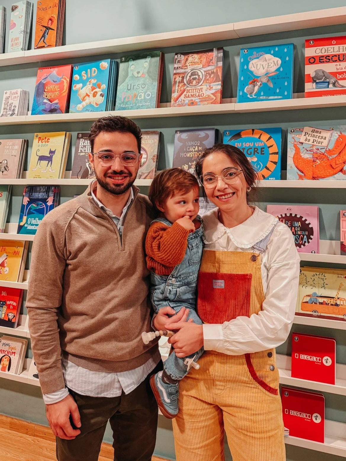 Família com um casal e uma criança posando para foto na livraria com prateleiras de livros ao fundo.