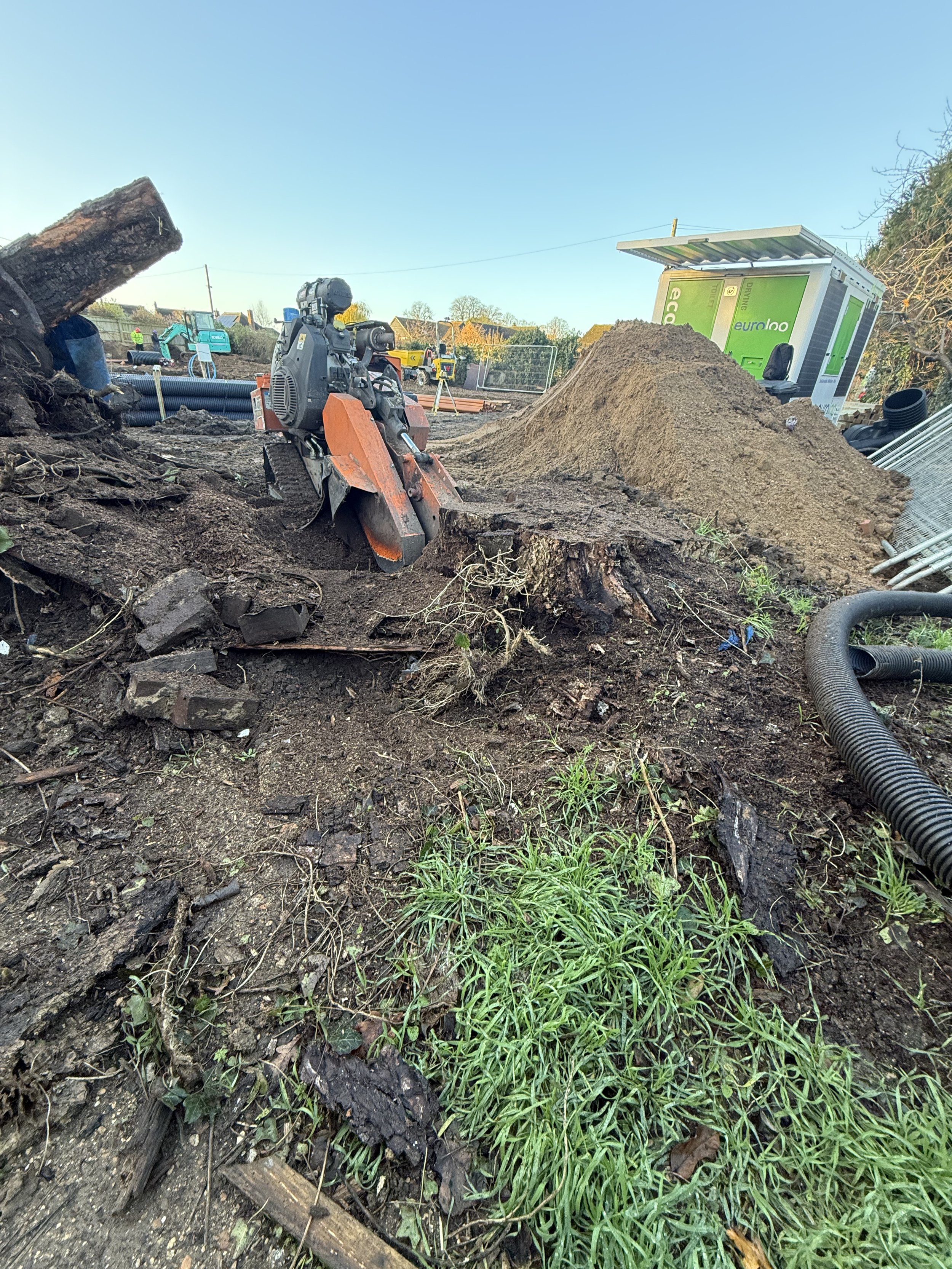 Construction site with dirt, a small excavator, a mound of dirt, construction materials, and a portable green unit in the background.