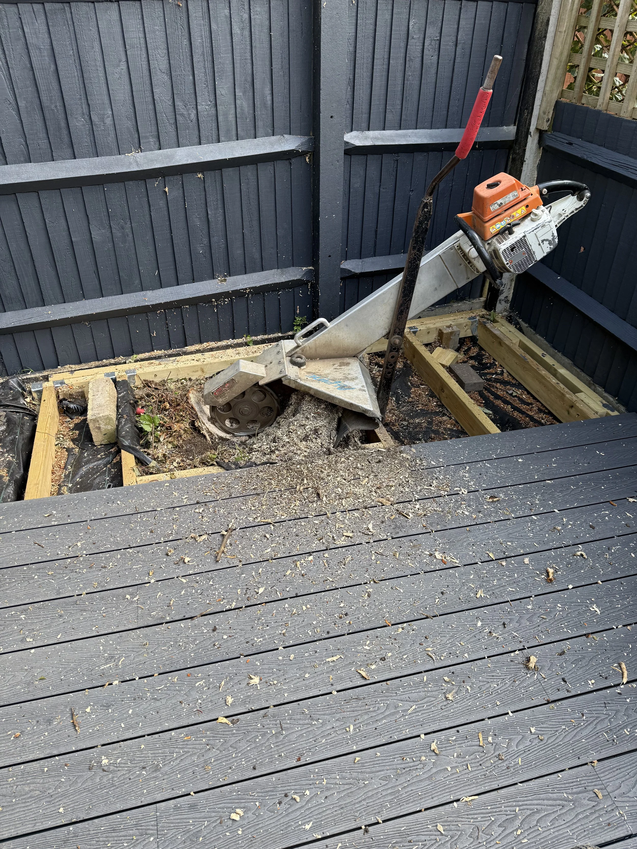 Construction site with a tree stump saw on a wooden deck, surrounded by wood framing and black painted fence panels.