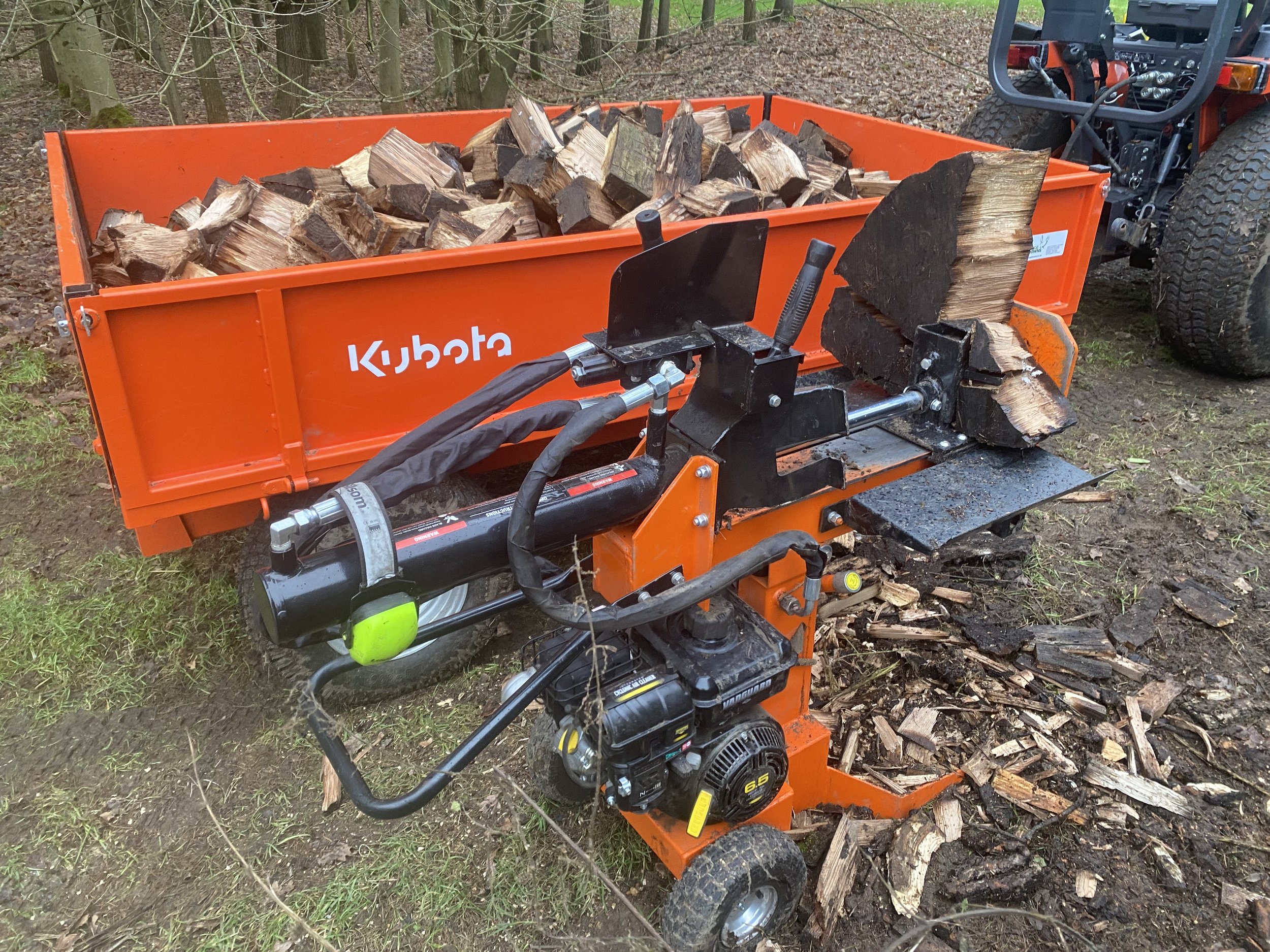 An orange wood splitter machine with the brand name 'Kubota' on the side, splitting logs into firewood, with chopped wood on the ground.