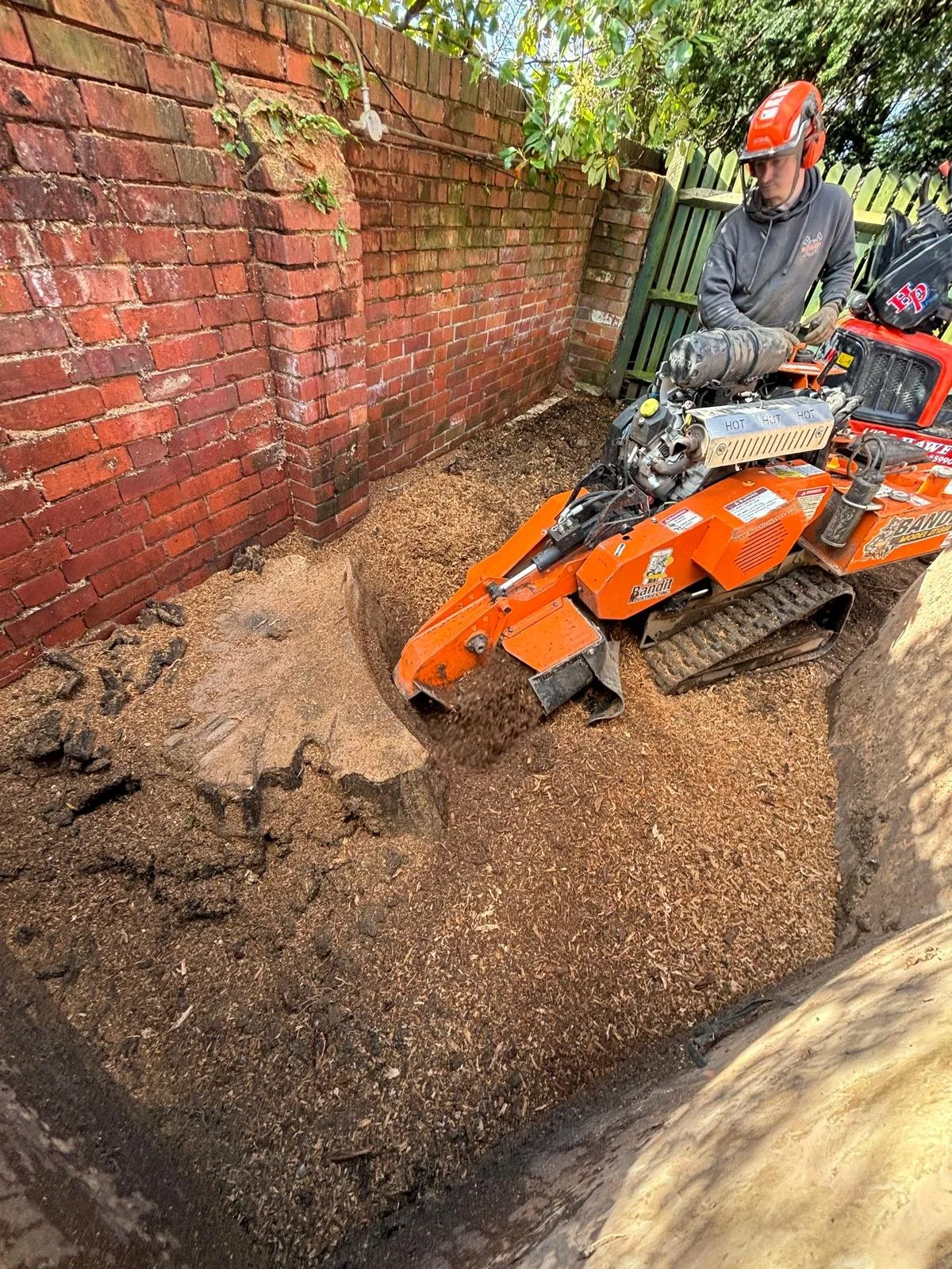A worker in a helmet using a small orange stump grinder to gradually remove a tree stump in a backyard with a brick wall and a wooden fence.