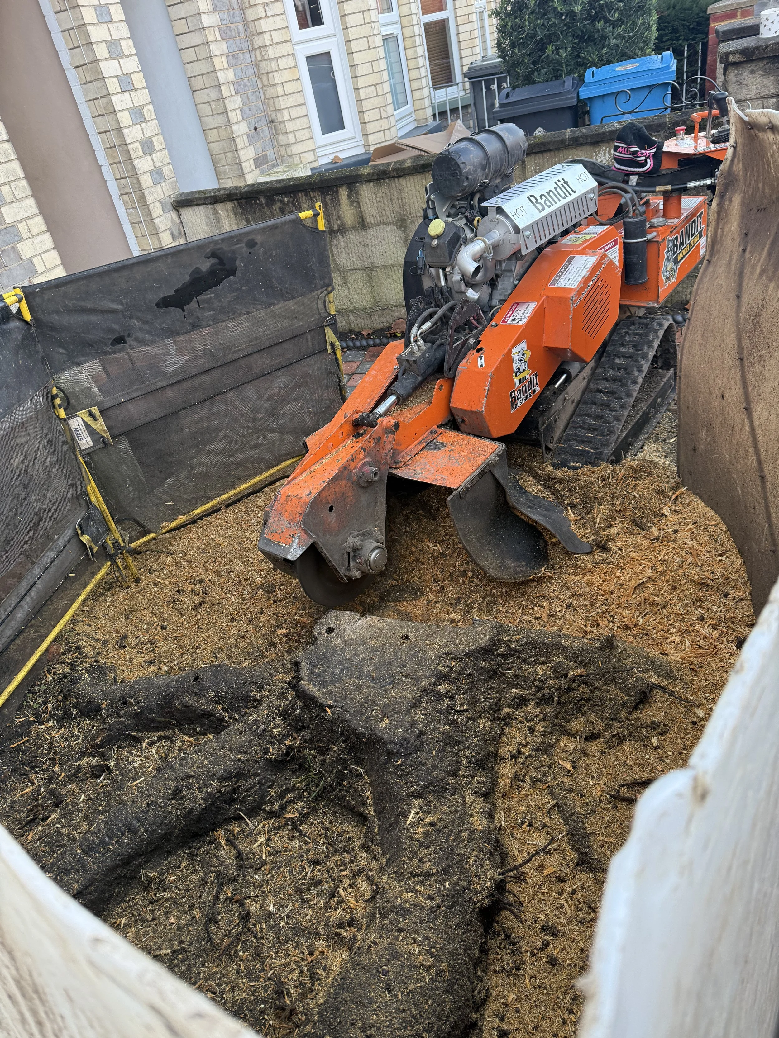 A small orange tracked construction or landscaping machine, removing a tree stump, on dirt and gravel, surrounded by temporary barriers on a residential street.