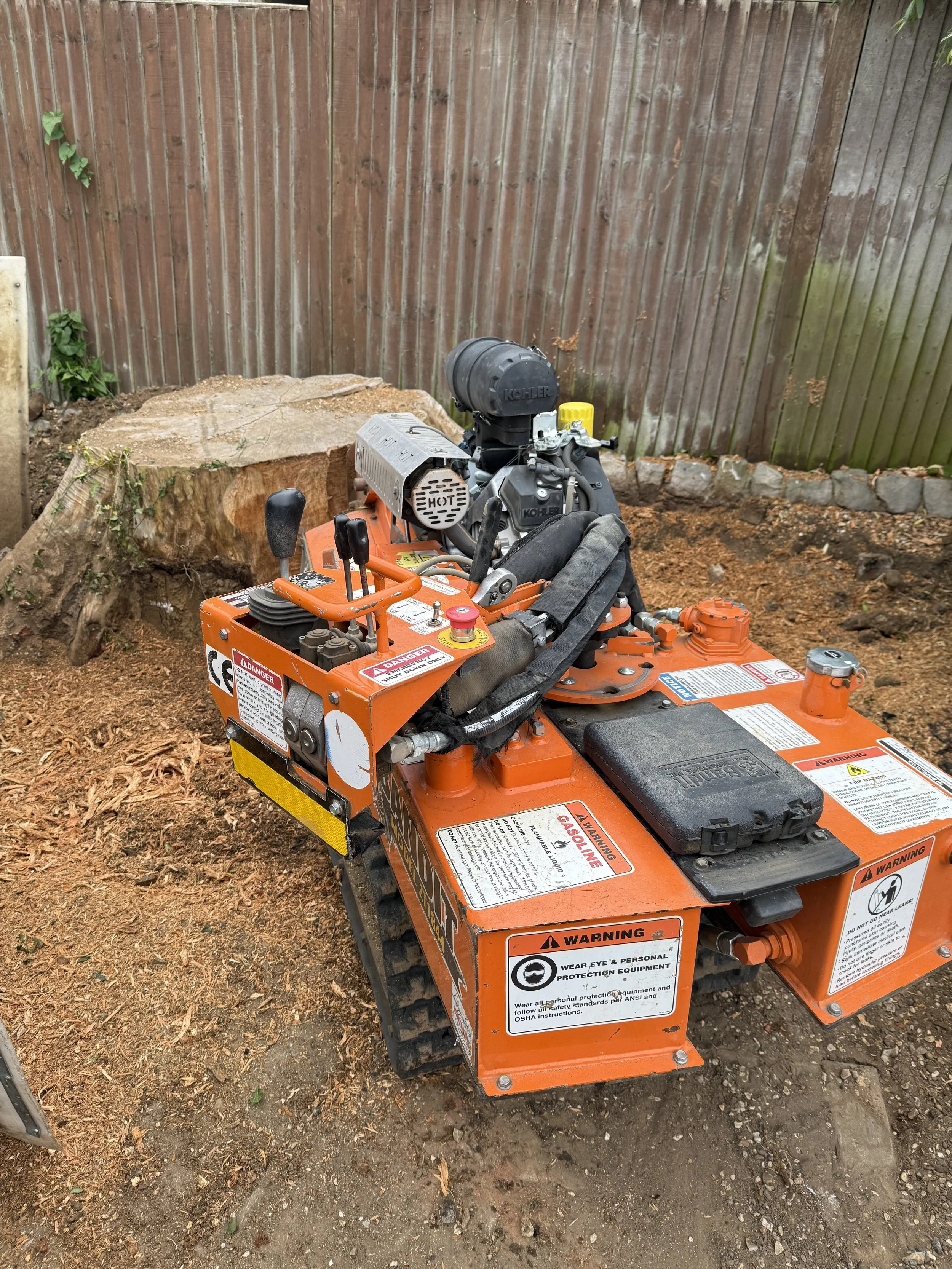A small orange tracked construction machine parked on a dirt surface with a large tree stump behind it and a wooden fence in the background.