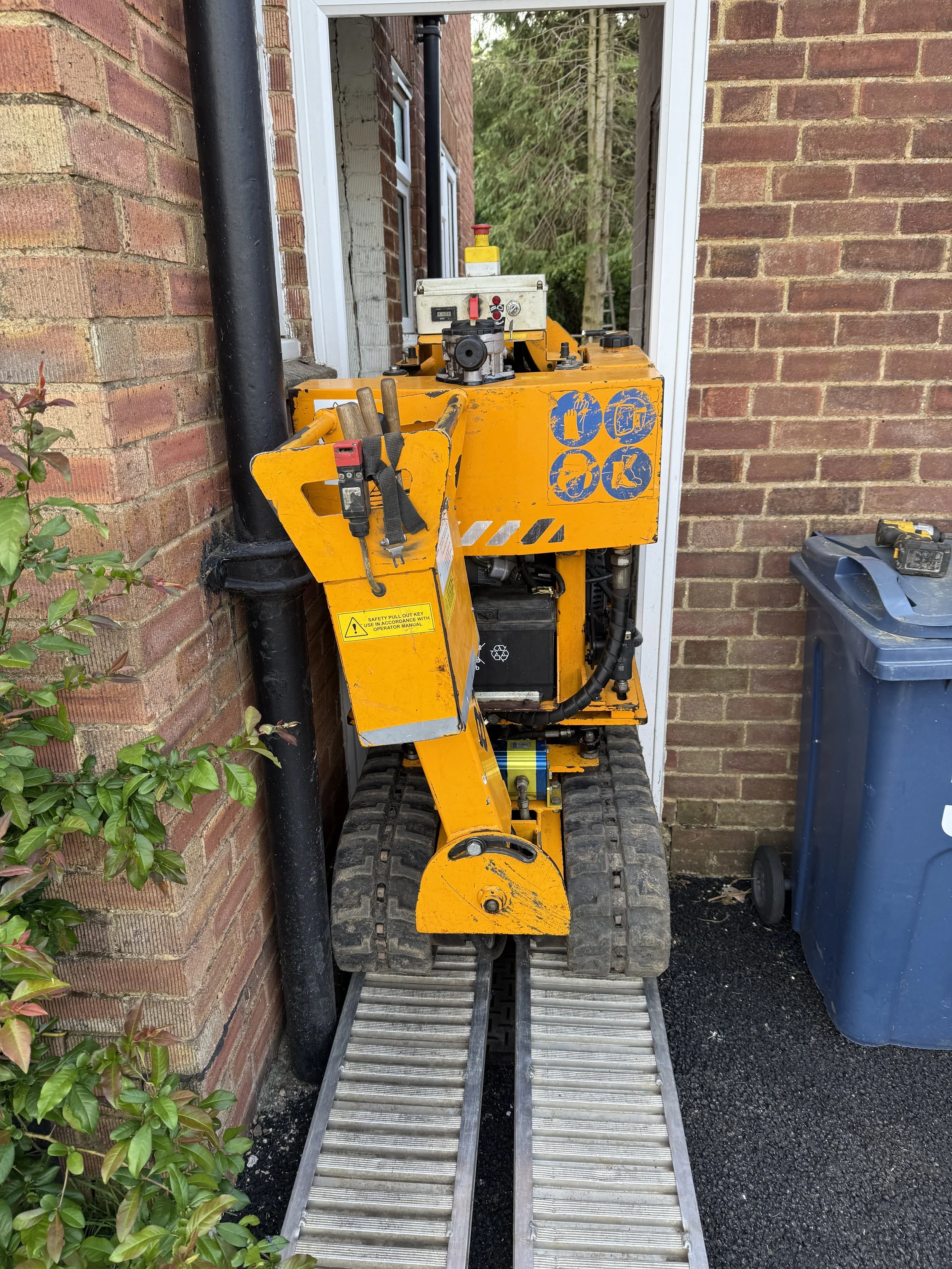 A small, yellow construction robot with tracked wheels, placed on a metal platform between brick walls, being brought through a  house side entrance, and next to a blue trash bin, outdoors