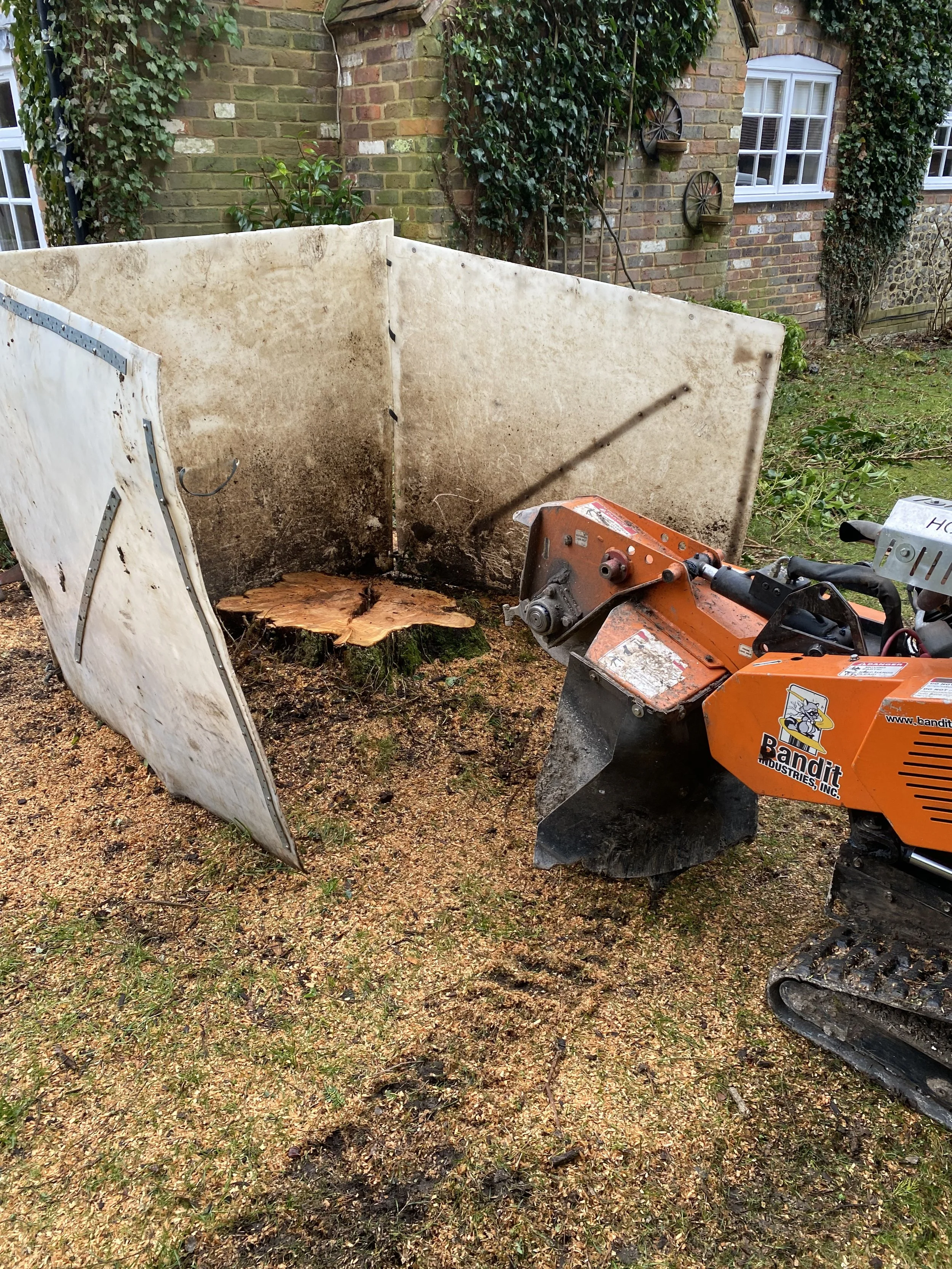 A small orange wood chipper machine in a yard, with a tree stump, a white protective barrier, and a brick house with ivy in the background.