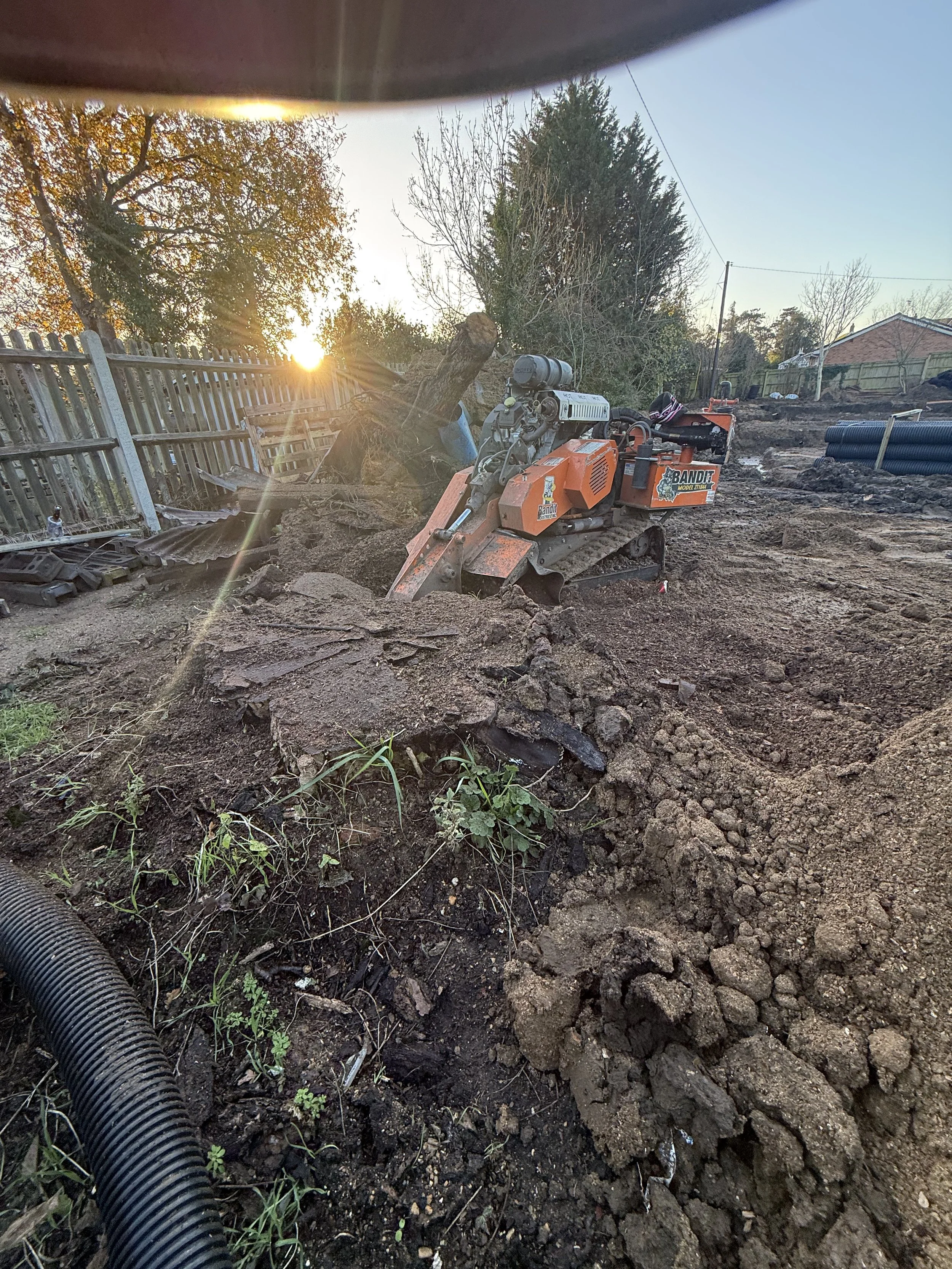 Construction site with an orange and black mini excavator working on digging soil, with a wooden fence and trees in the background, and the sun setting in the distance.