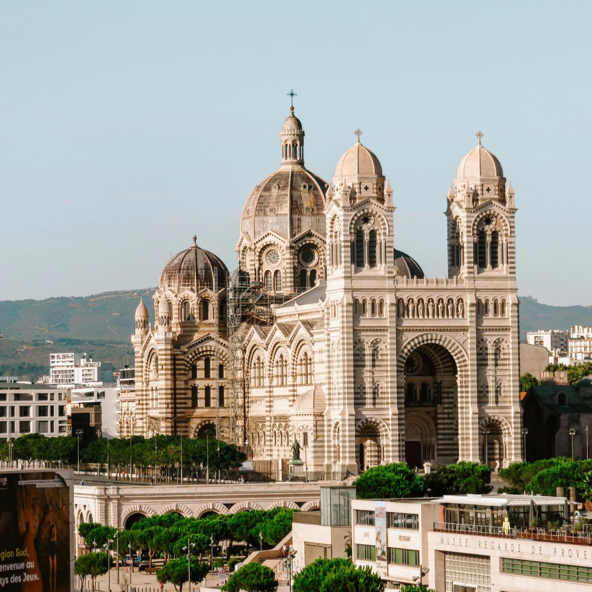 A large, ornate cathedral with striped stonework, multiple domes, and twin bell towers, situated in an urban area with trees and modern buildings in the foreground, and hills in the background.