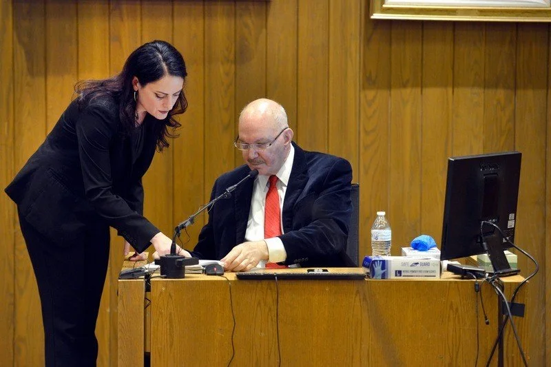 A woman is leaning over a man sitting at a desk in a wood-paneled room, possibly in a courtroom or meeting room. The man is looking at documents, while the woman appears to be discussing or pointing to the papers.