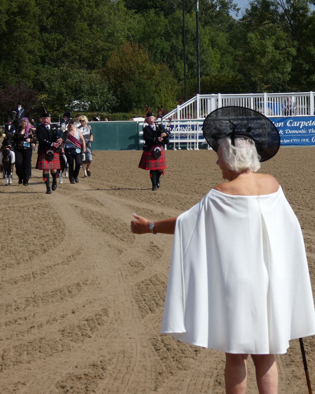 An elderly woman in a white dress and large black hat stands on sandy ground, watching a parade of bagpipers and others in formal attire, some on horseback, at an outdoor event with trees in the background.