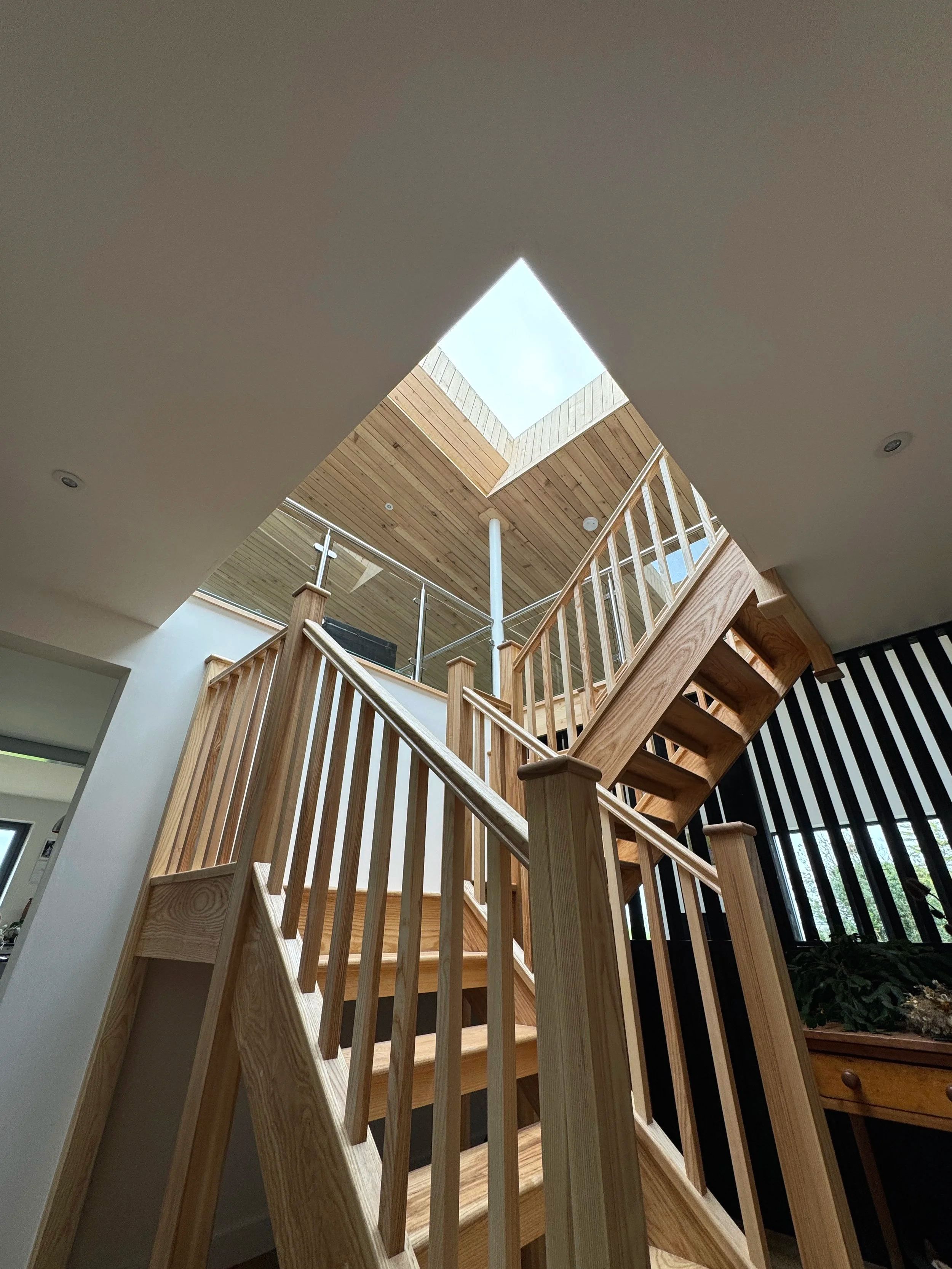 Interior view of a wooden staircase with railings, leading up to a skylight ceiling in a modern home.