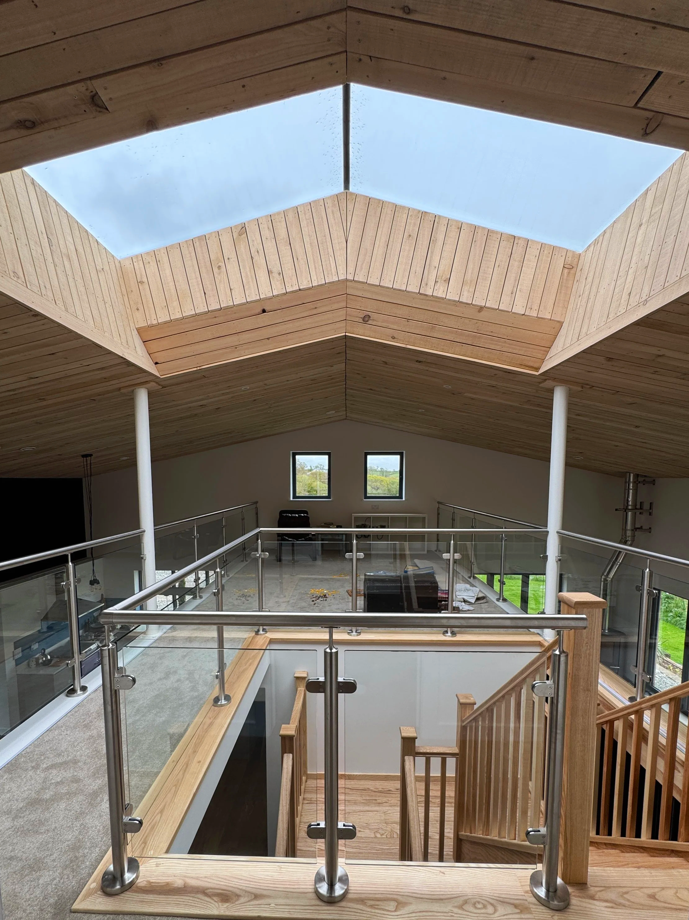 Interior view of a spacious, modern house with wooden ceiling and a large skylight, glass railings, wooden staircase.