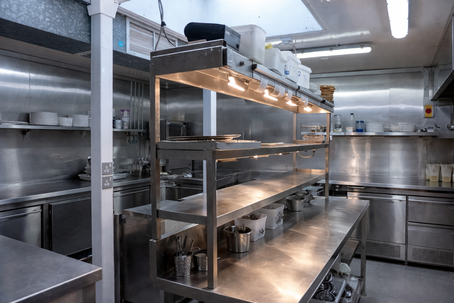 Empty stainless steel commercial kitchen with shelving and counters, some dishes and utensils on shelves, and overhead lighting.