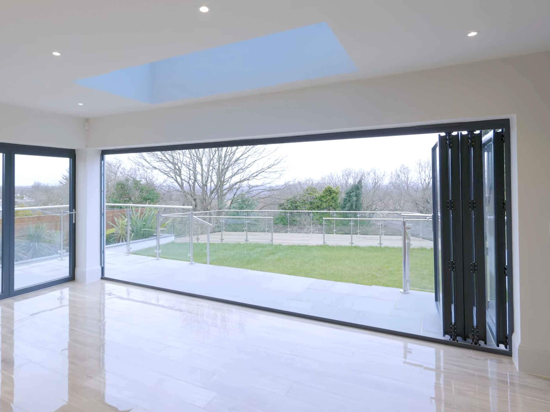 Modern living room with large sliding glass doors opening to a backyard with grass, trees, and a paved patio, under a ceiling with a skylight.