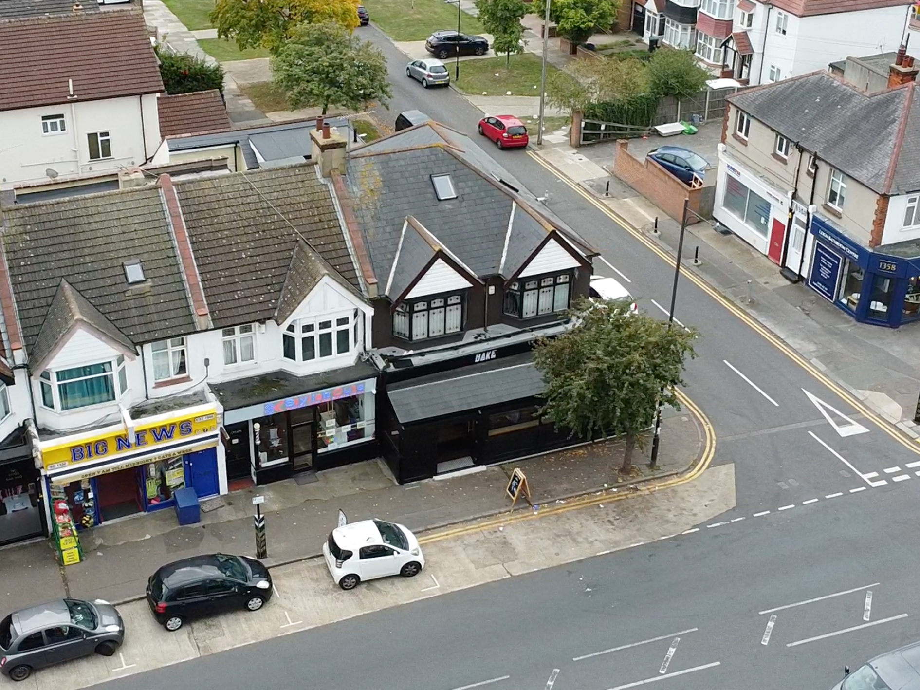 An aerial view of a residential street showing several small shops with storefronts, parked cars on the street and sidewalk, houses with tiled roofs, a small tree, and a crosswalk at the corner.