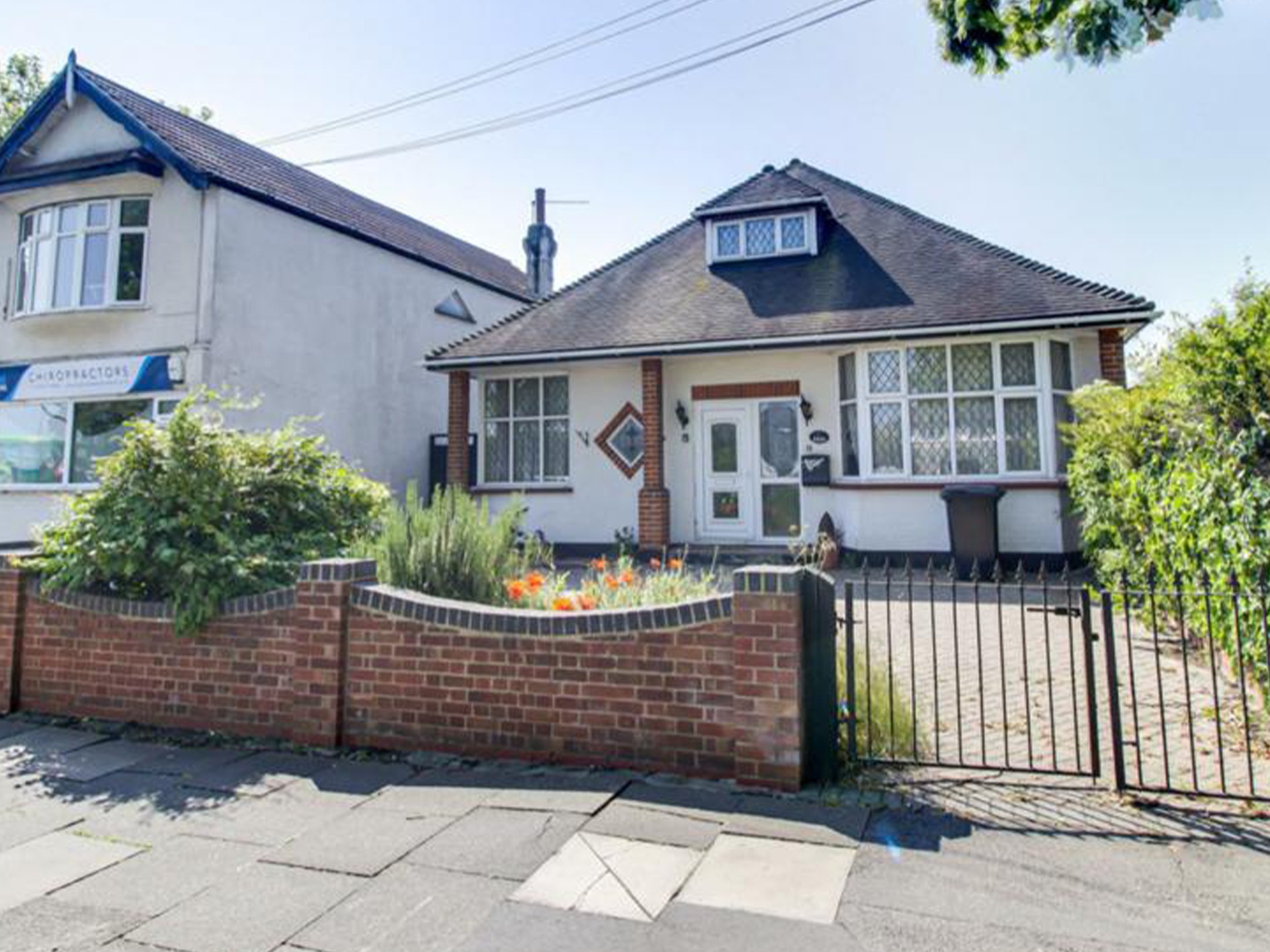 A house with a white exterior, brick accents, and multiple windows, surrounded by a small garden and a brick and metal fence, under a clear blue sky.