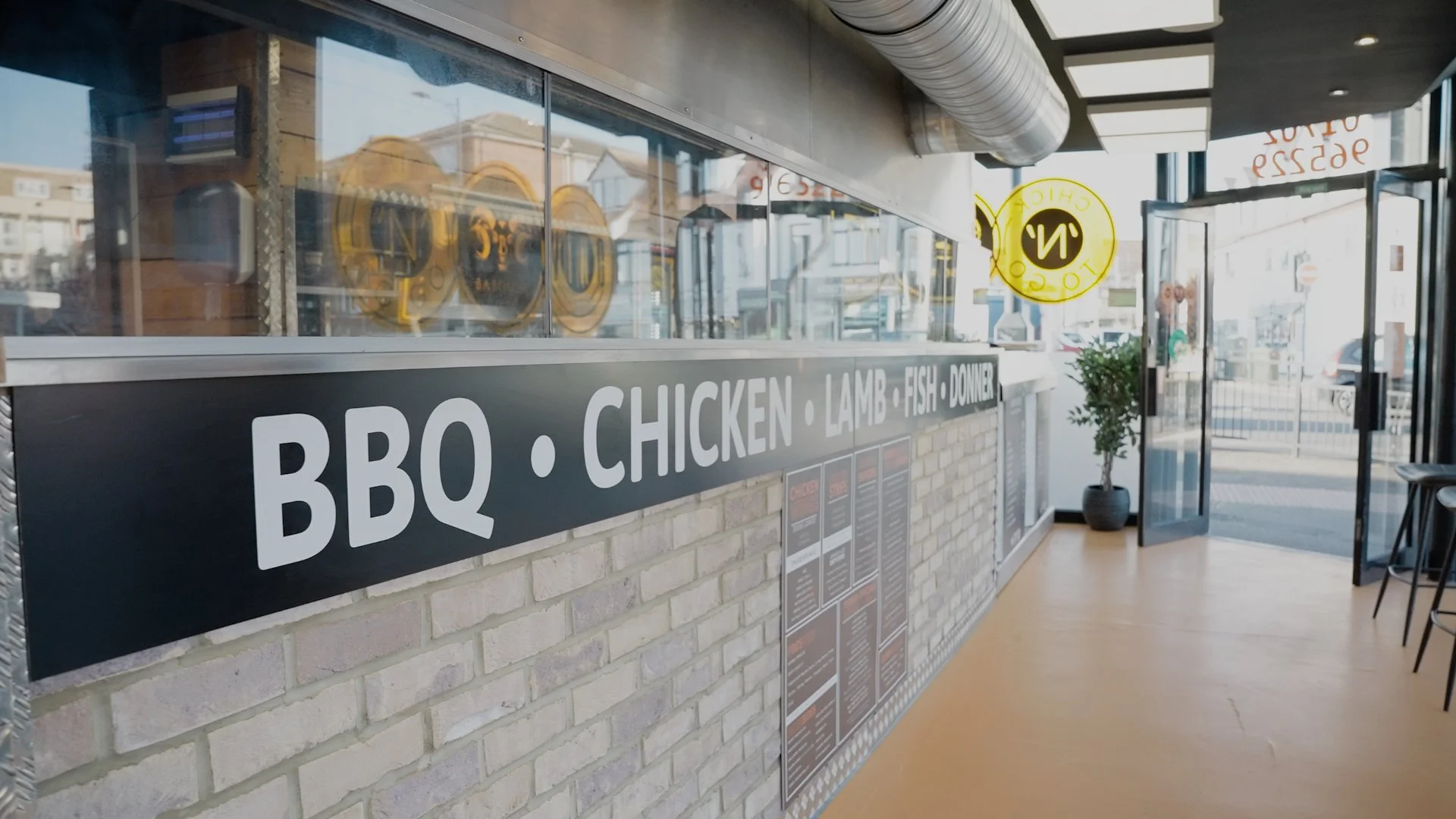 Inside a fast food restaurant with a black sign displaying 'BBQ, Chicken, Lamb, Fish, Doner'. The interior includes a brick wall, a menu, a door, and a potted plant near the entrance.