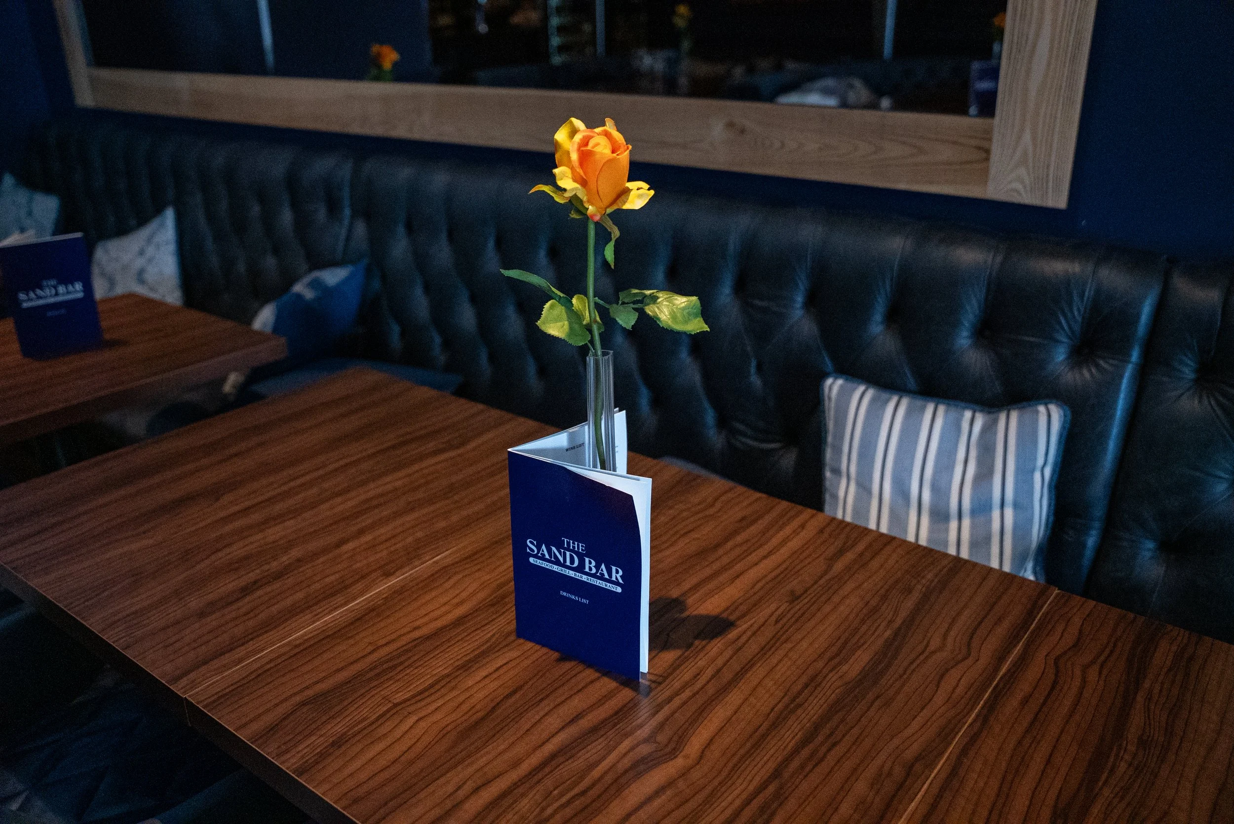 A table with a blue menu and a single orange rose in a tall clear vase, placed on the menu, inside a restaurant or bar called The Sand Bar.