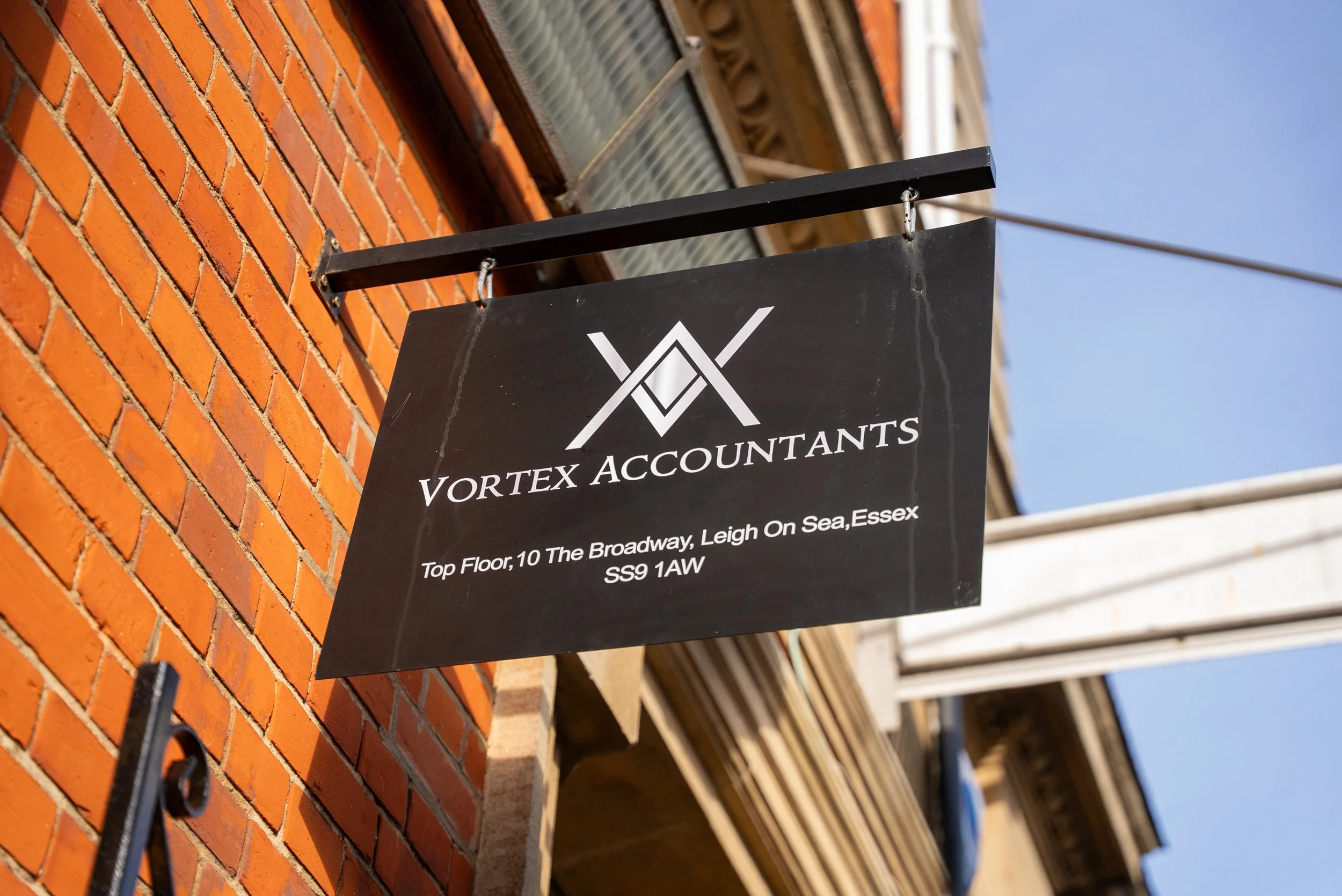 Black signboard for Vortex Accountants hanging outside a brick building with a clear blue sky in the background. The sign displays the company logo and address in white text.