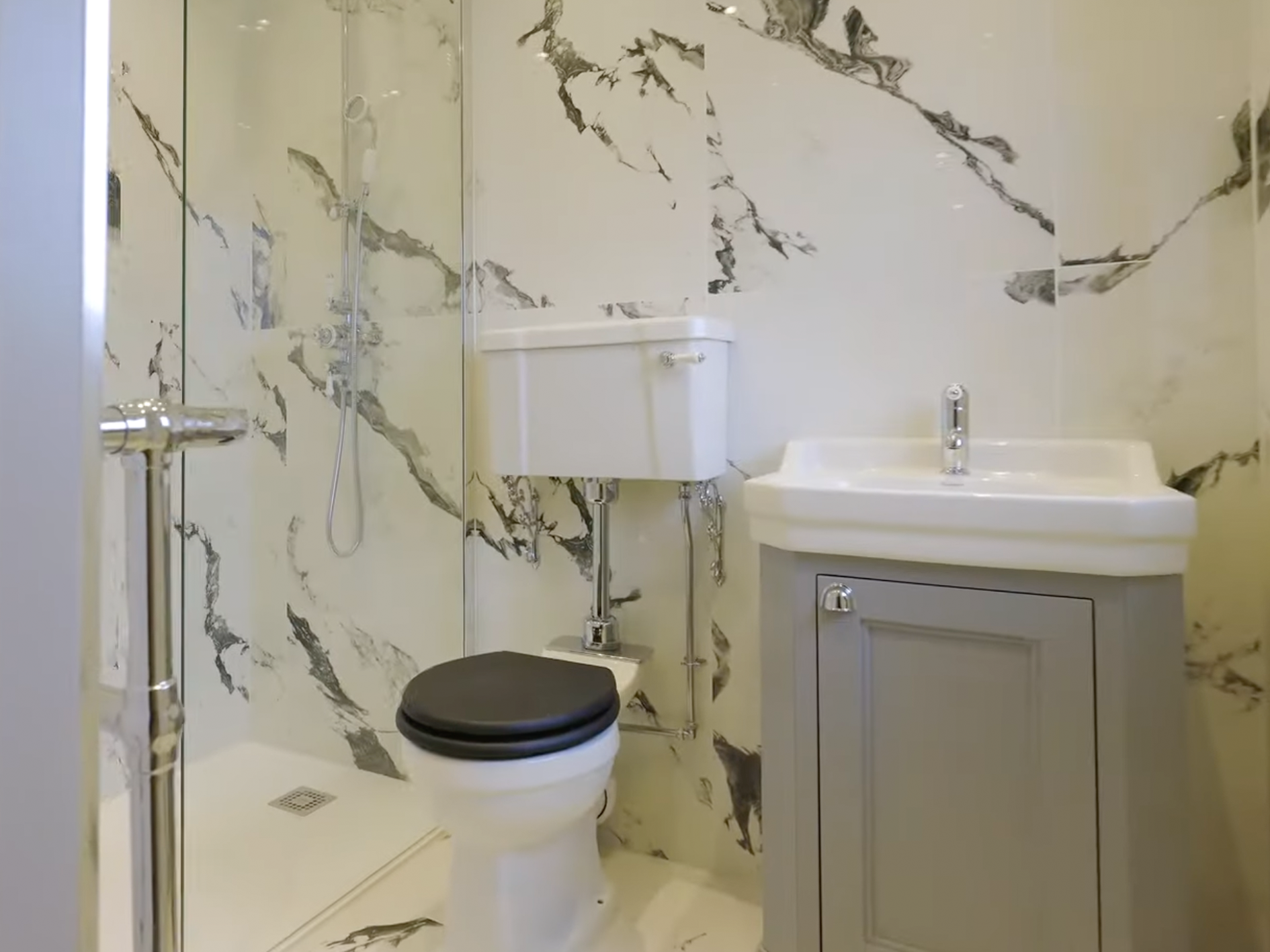 Small bathroom with marble-patterned walls, a compact shower area with a handheld showerhead, a toilet with a black seat cover, and a white sink with a gray cabinet beneath.