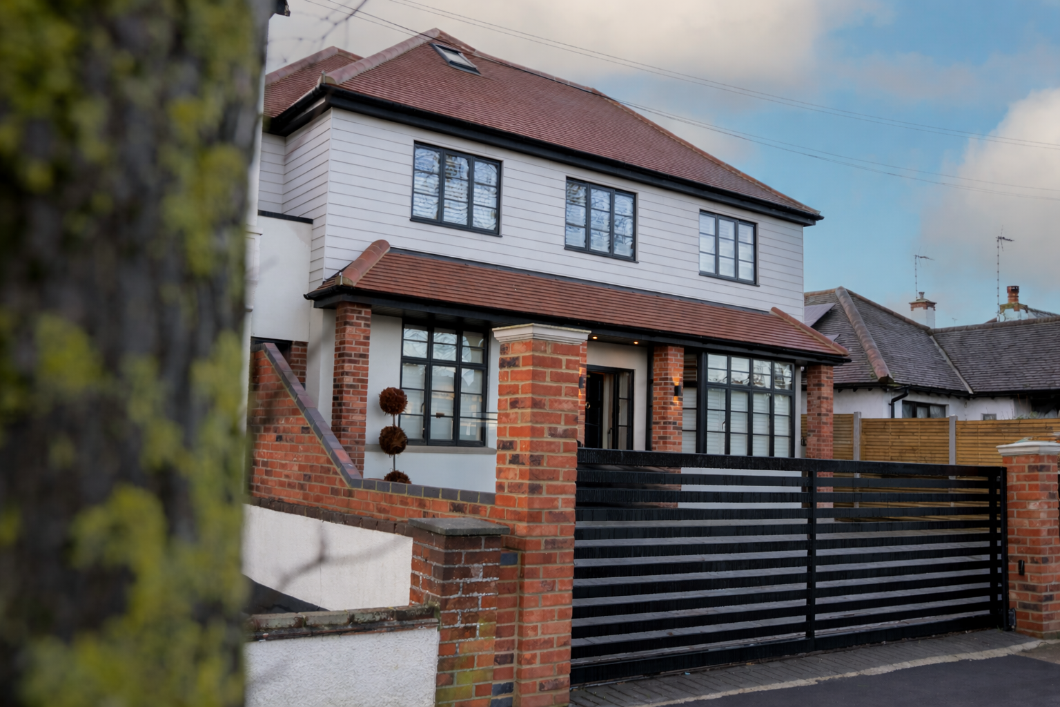A modern two-story house with a brick and white exterior, black window frames, and a red tiled roof, set behind a black wooden gate and brick fence.