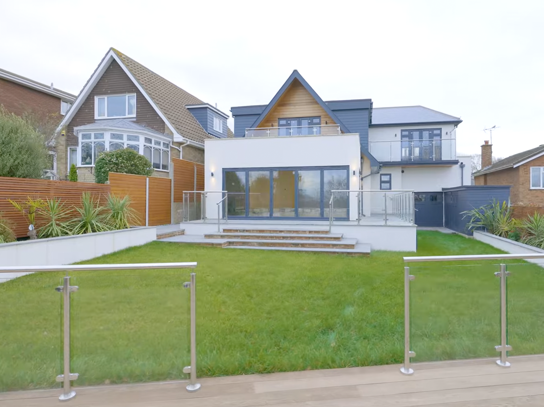 Modern house with large glass sliding doors, white exterior, steps leading to a green lawn, and neighboring homes with gardens.