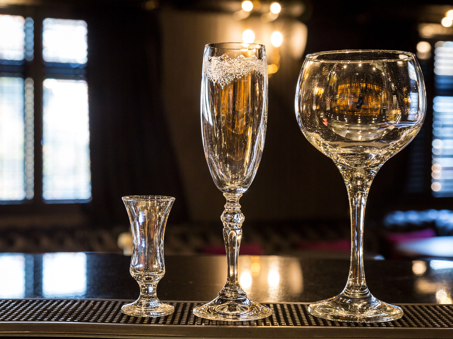 Four empty glasses of different shapes on a bar counter in a dimly lit room with windows in the background.