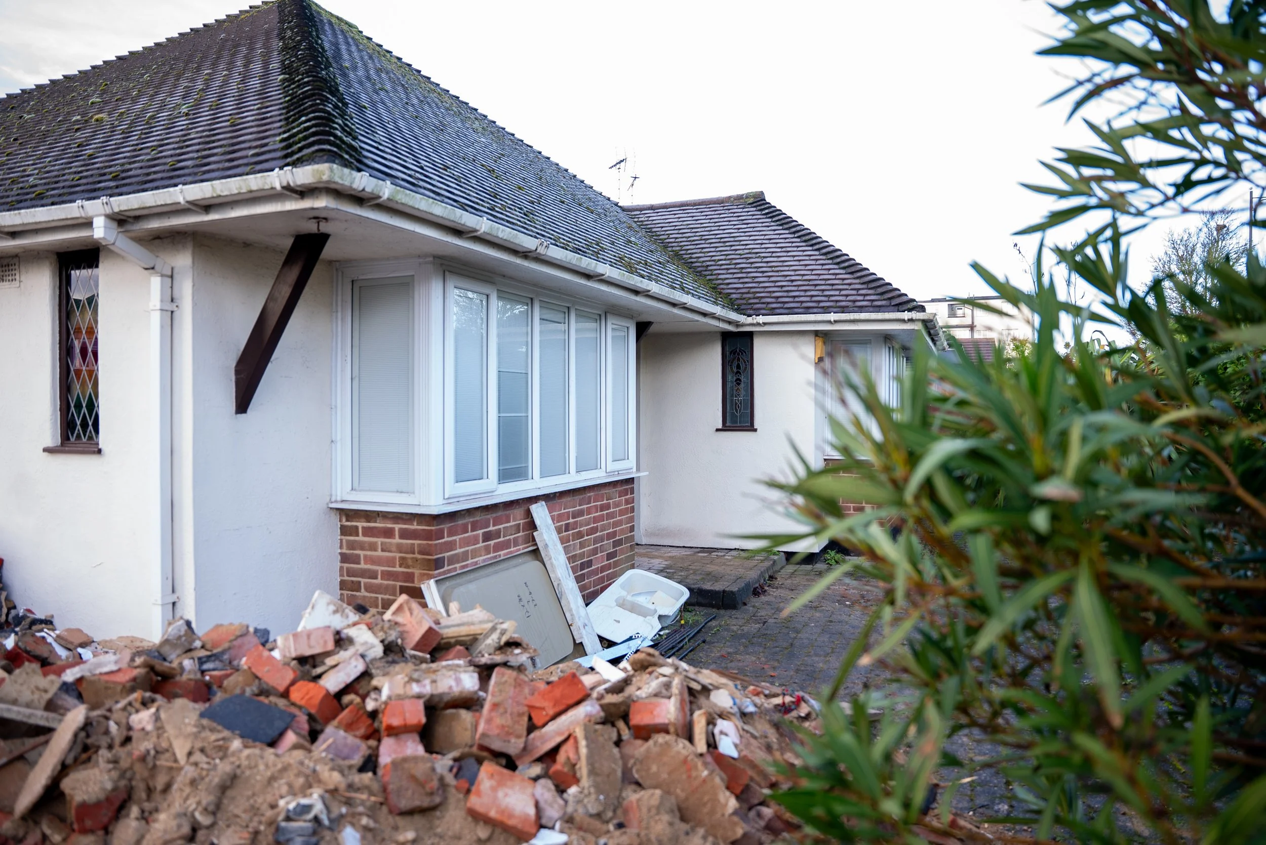 A house with a white exterior, brick foundation, and a tiled roof. There is a pile of bricks and construction debris in the foreground, and some green shrubbery on the right side.