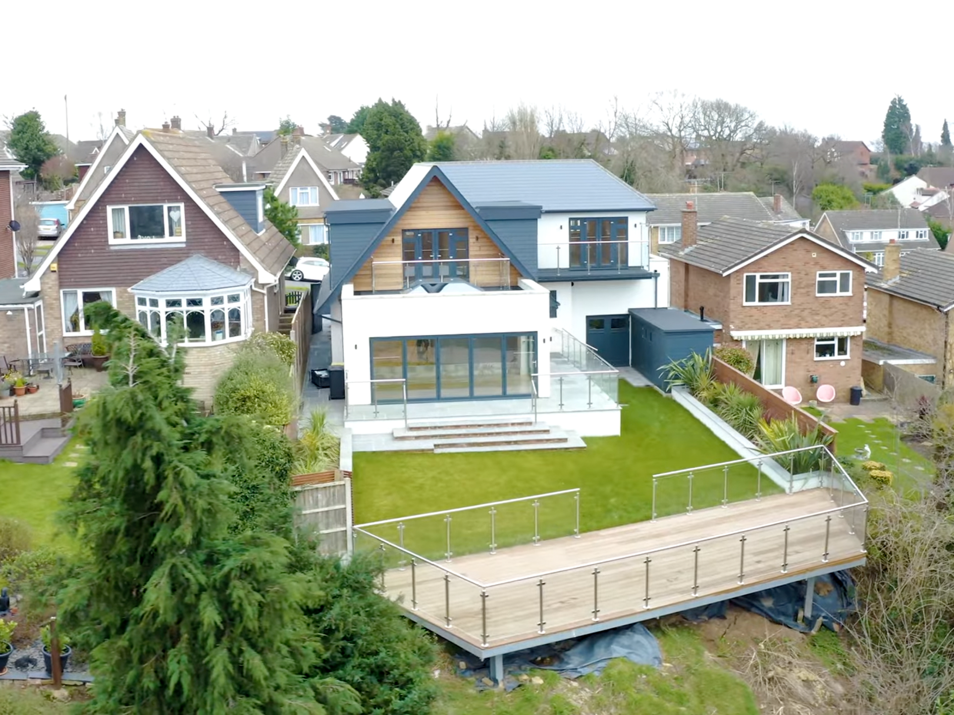 A modern house with white exterior walls, large glass doors and a spacious wooden deck with glass railing in the backyard, surrounded by neighboring suburban houses and greenery.