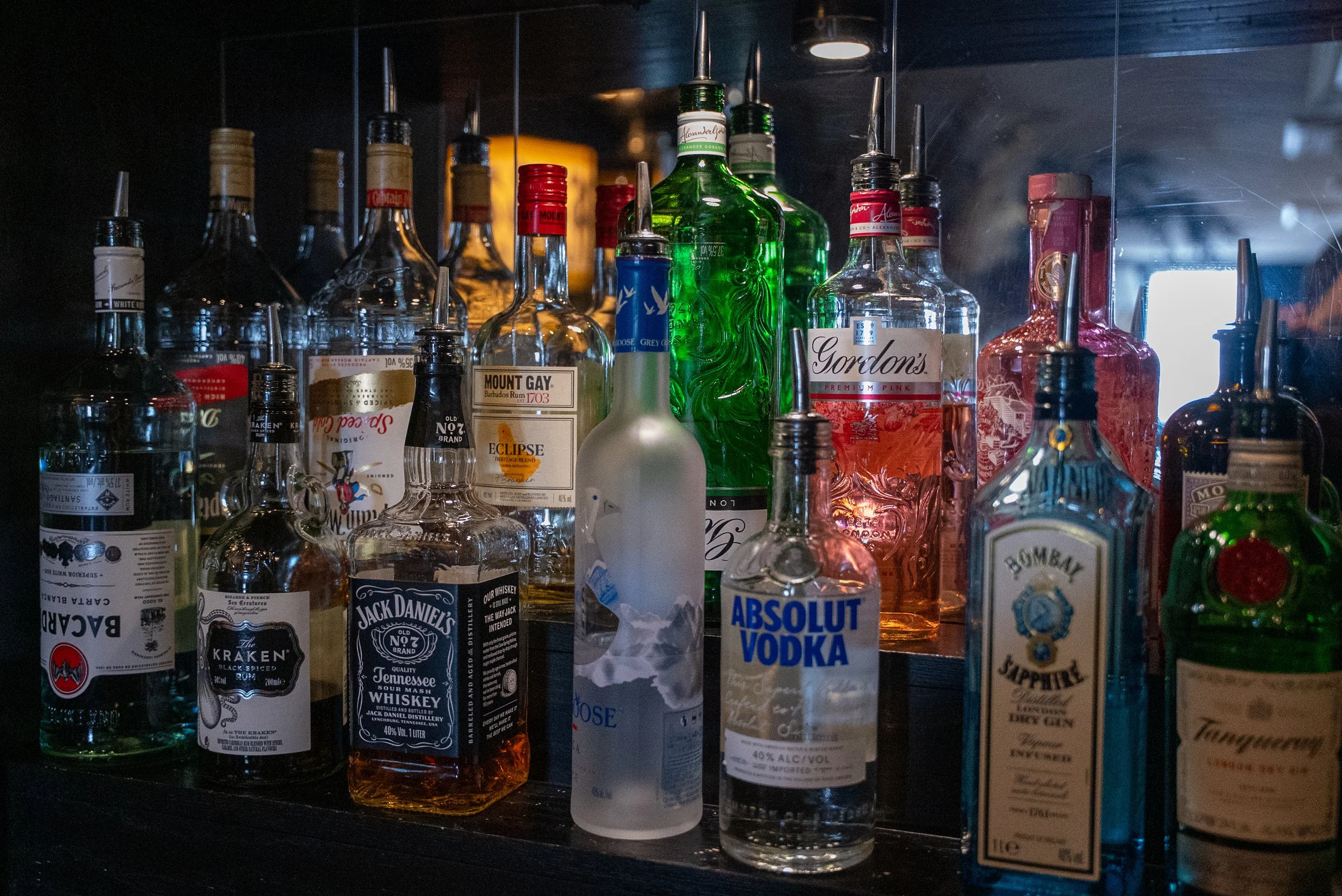 A collection of various liquor bottles, including Jack Daniel's whiskey, Absolut vodka, Bombay Sapphire gin, Gordon's gin, Campari, Tanqueray gin, and several others, arranged on a dark shelf or bar.