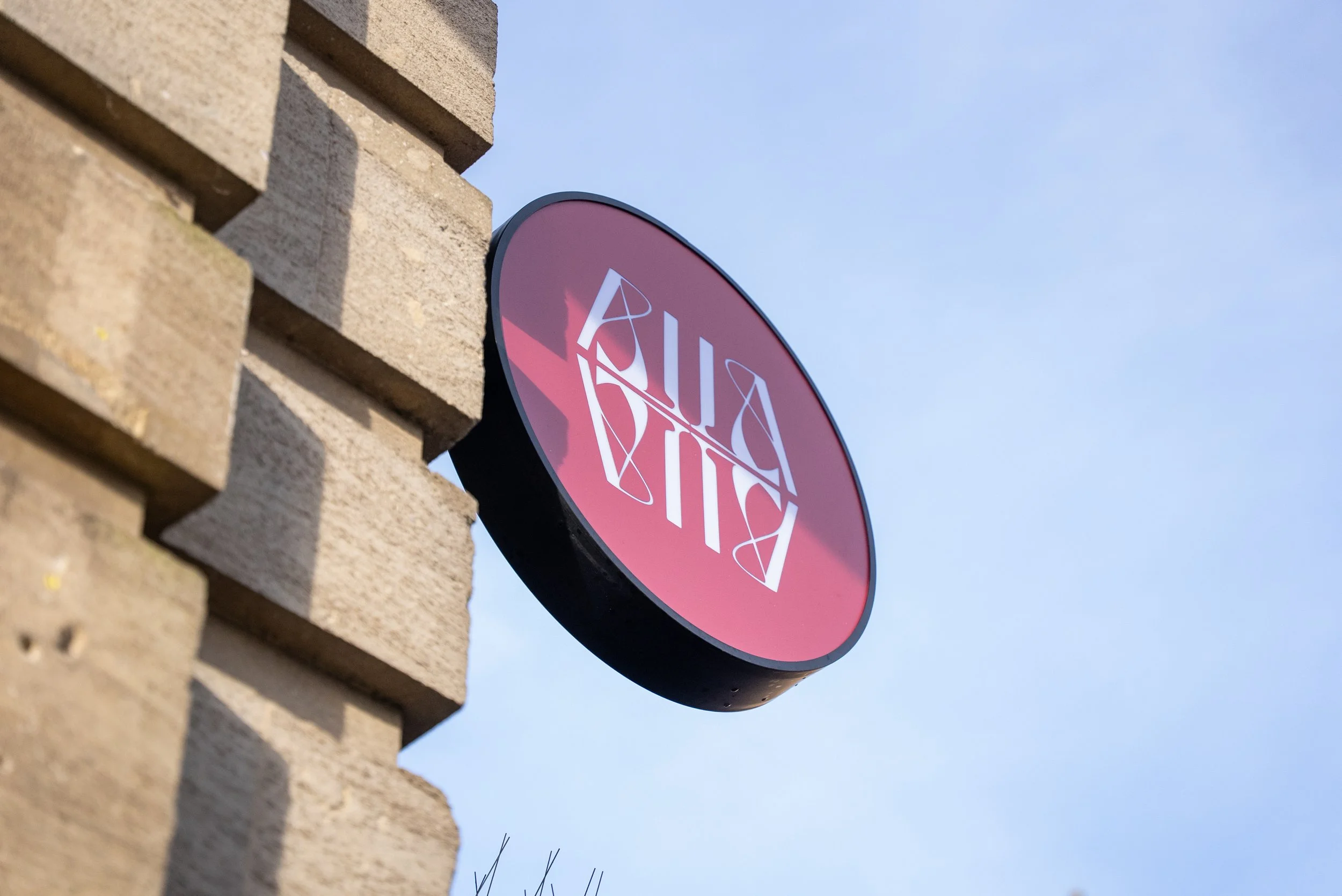 A round pink sign with white text that says "LA LA Lagree" against a clear blue sky, attached to a brick building.