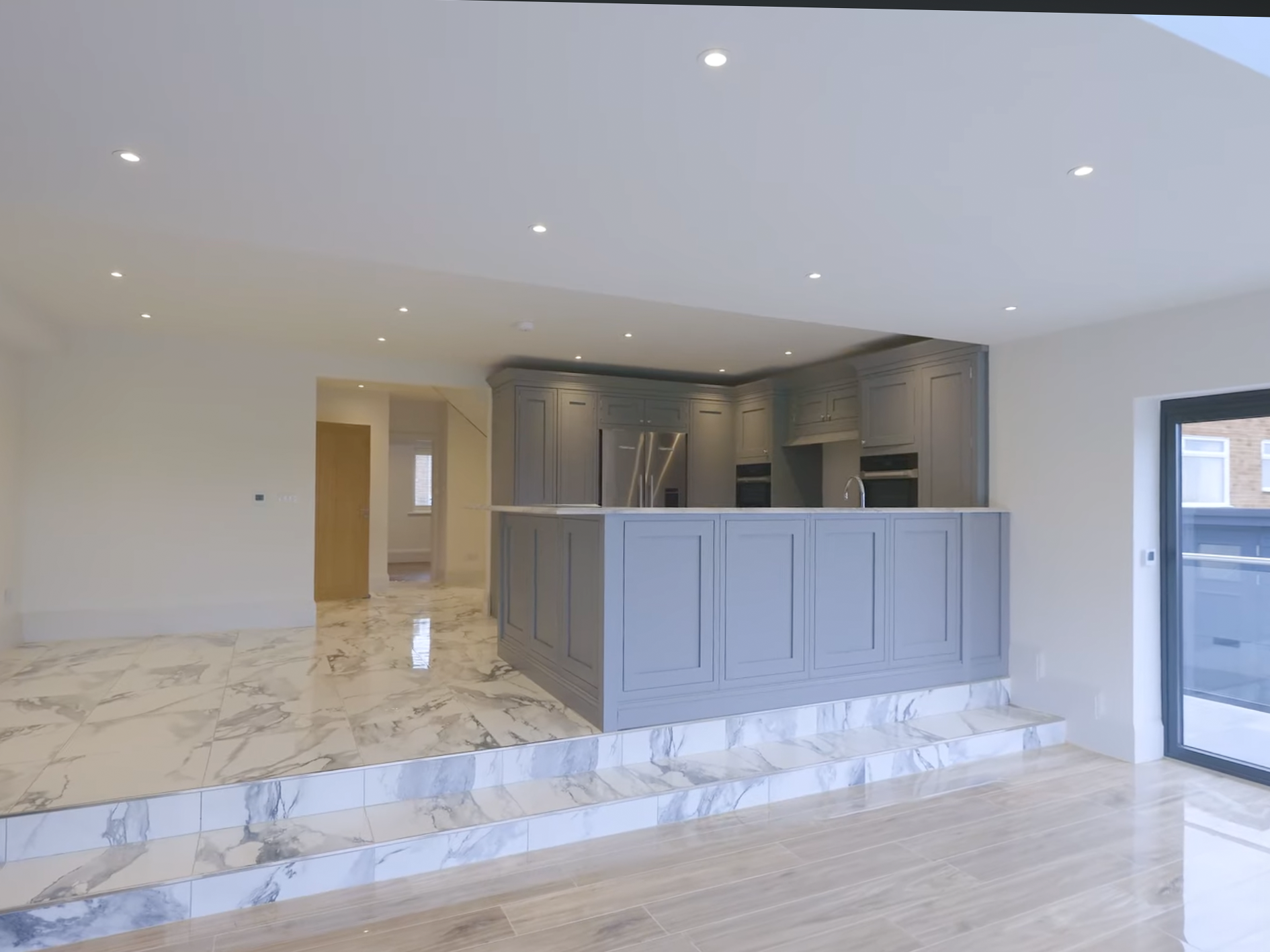 Modern open-plan kitchen with gray cabinetry, marble floors, and a large window or sliding door, with recessed ceiling lights.