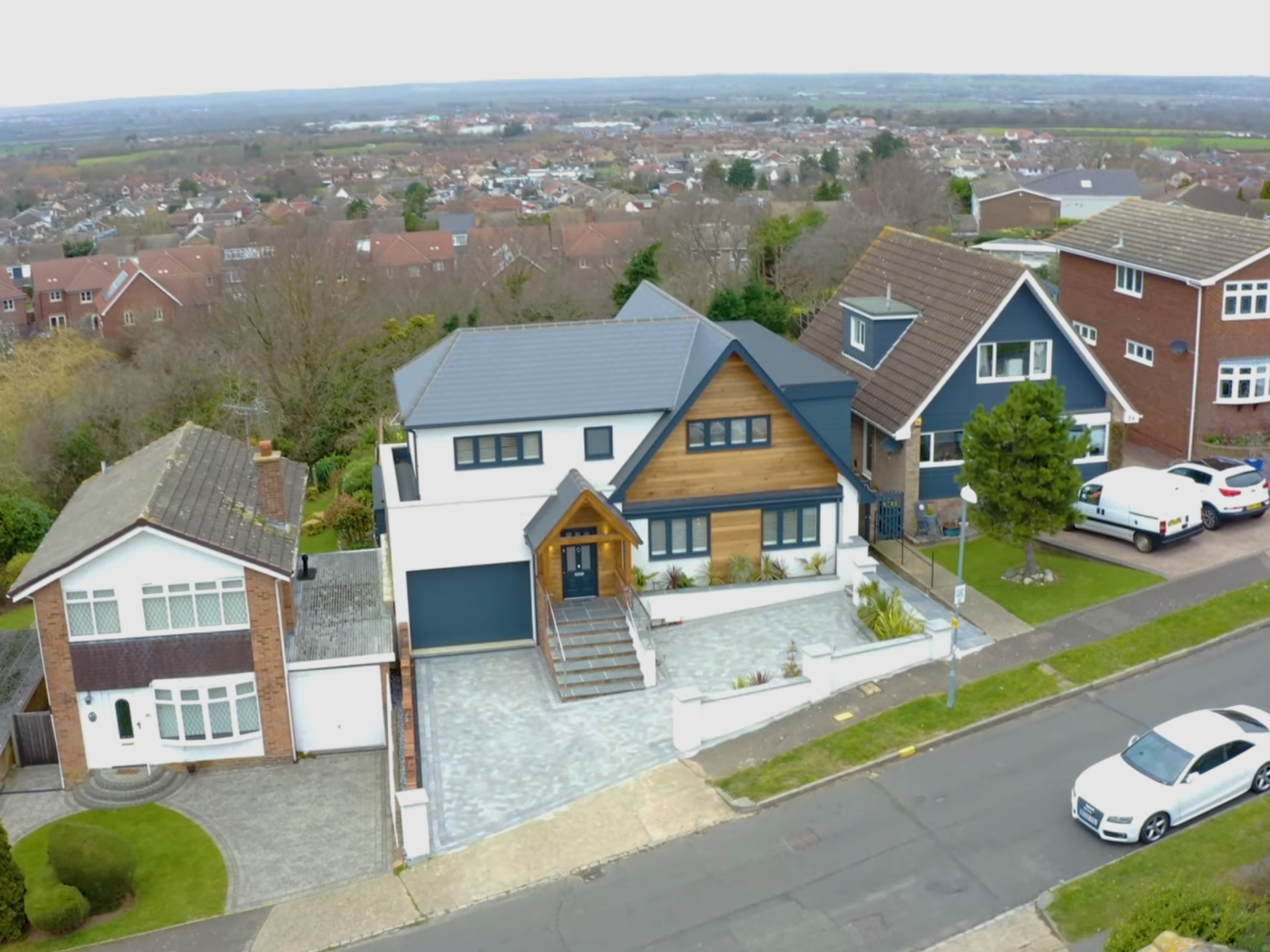 Aerial view of a suburban neighborhood showing three houses, a park area with a lamp post, cars parked along the street, and more houses in the distance.