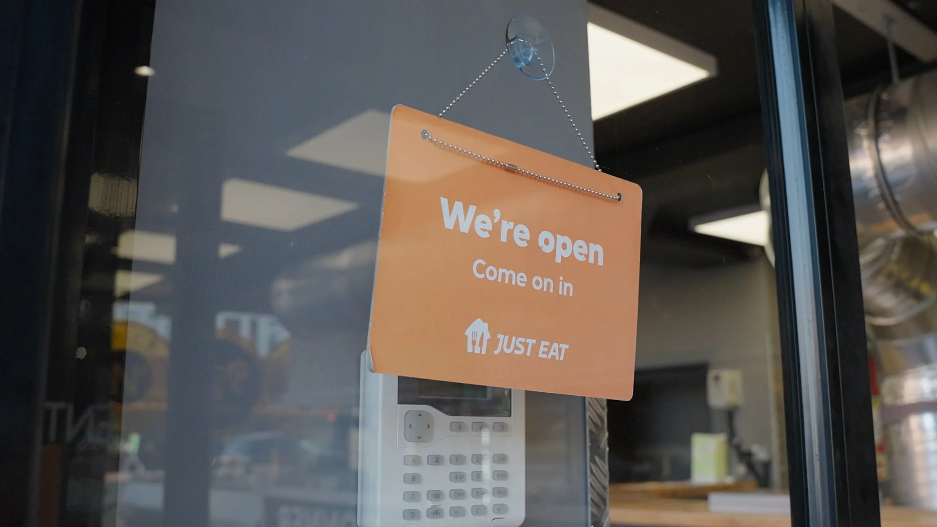 Orange sign on glass door that says "We're open. Come on in" with the Just Eat logo below, and a white intercom or phone visible behind the glass.