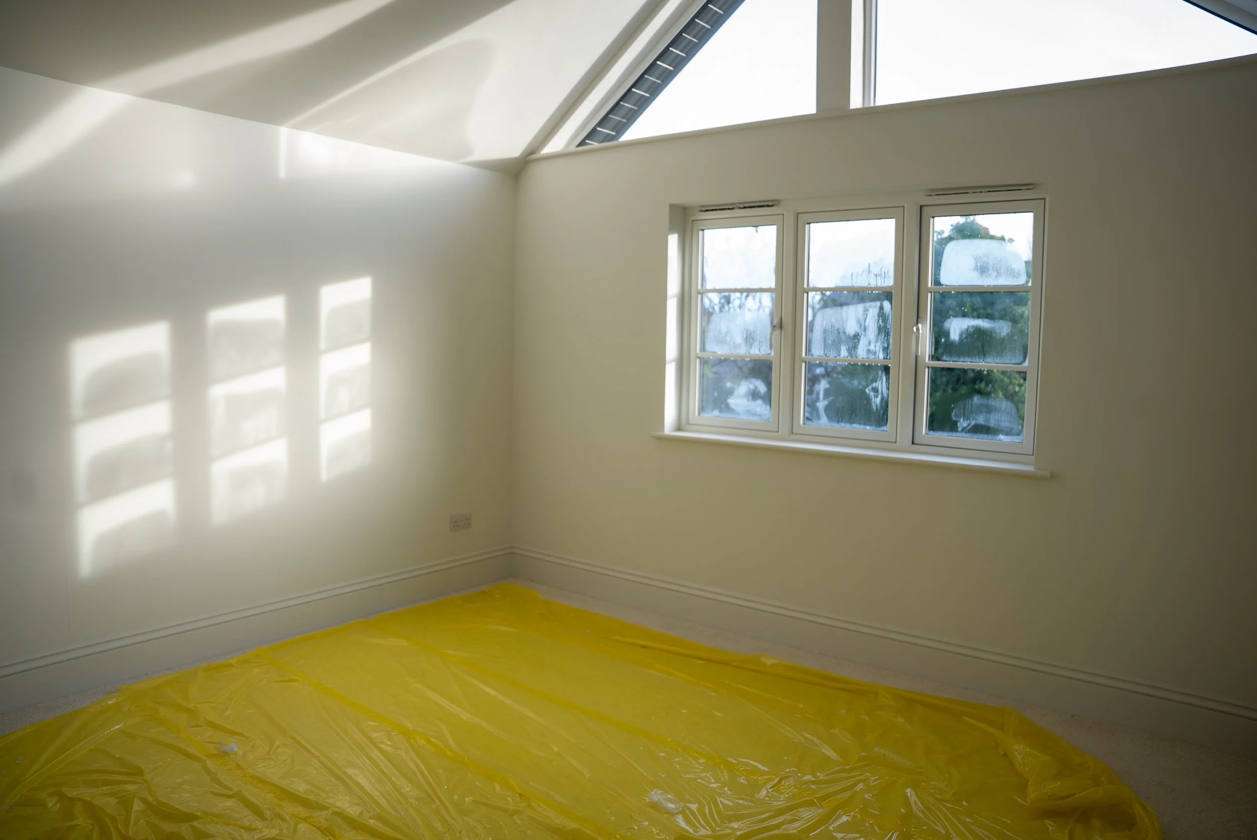 Empty room with white walls, a large window with condensation on the glass, and a yellow plastic sheet on the floor, possibly for painting or renovation.