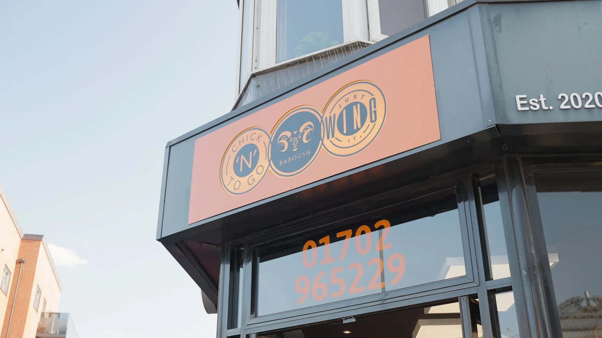 Sign on a building with a logo reading "BABOUSH" and the words "CHICKEN TO GO" arranged in circles. The sign has a peach-colored background and is located above a glass door with a partial view of a phone number in orange. The building has a gray exterior and the establishment was established in 2020.
