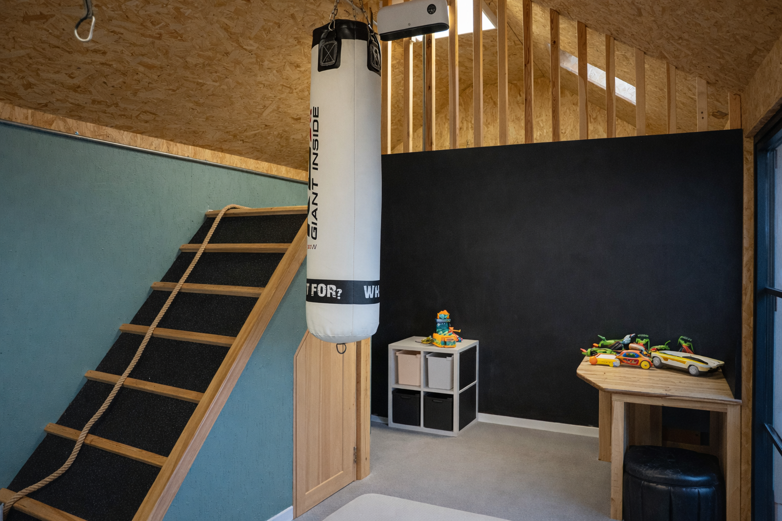 Children's playroom with a boxing punching bag hanging from the ceiling, a climbing ladder with rope, a small shelving unit with storage bins, and a wooden table with toy guns and cars on it, next to a black wall.