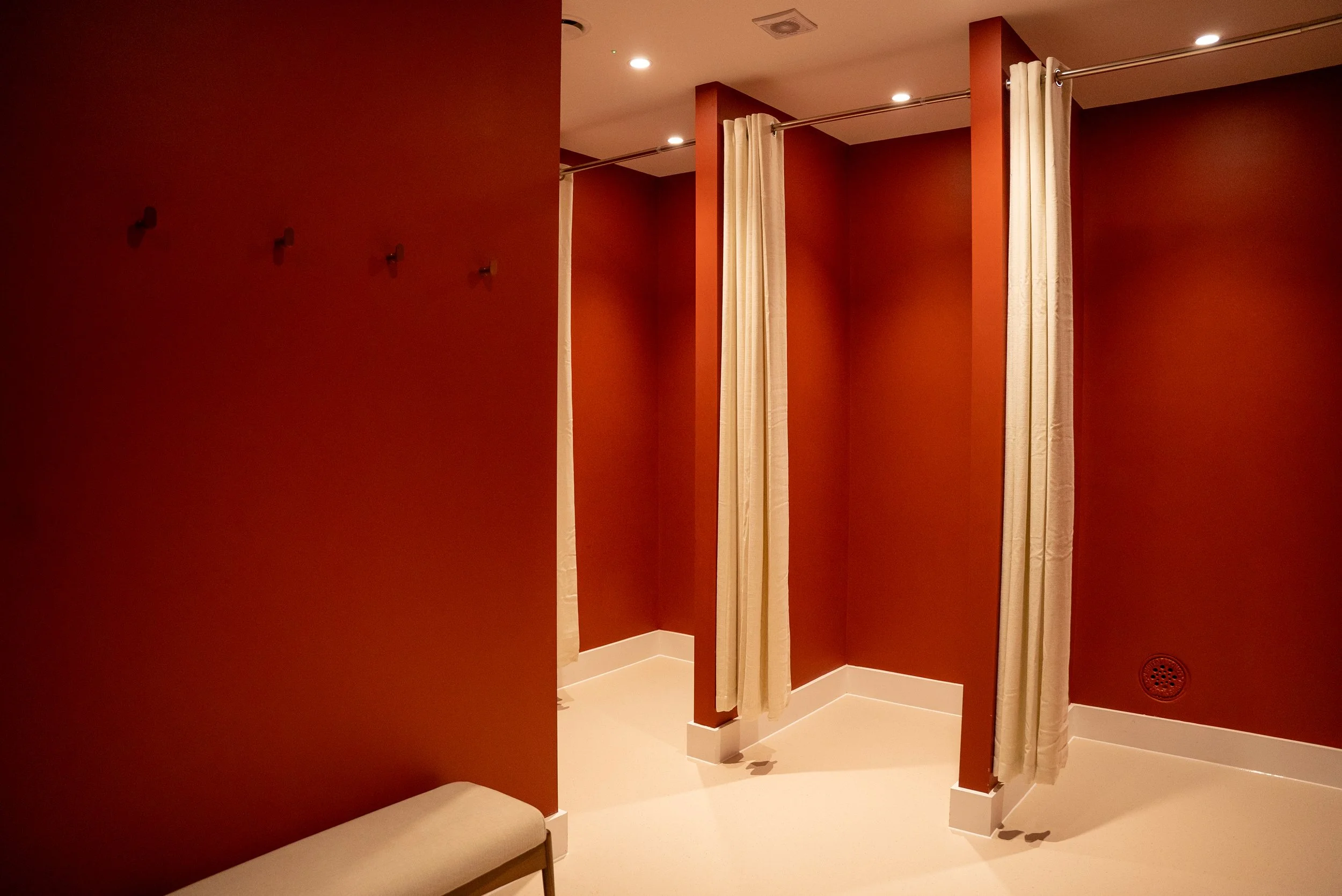 Public changing room with beige curtains dividing sections, rust-colored walls, white trim, and a beige bench.