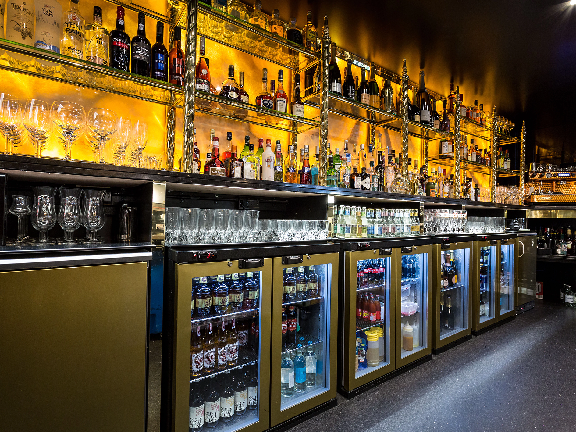 Bar with shelves of liquor bottles, glassware, and mini fridge units, illuminated with warm yellow lighting.